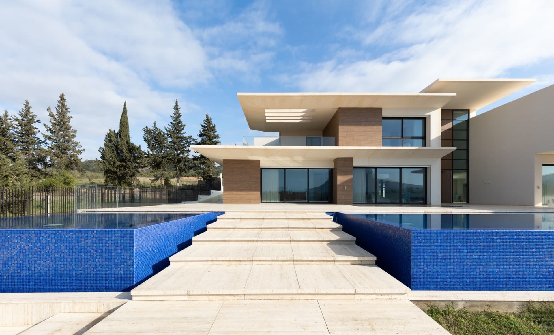 Modern residential exterior featuring a pool with blue tiled edges, surrounded by concrete and glass materials, under a clear