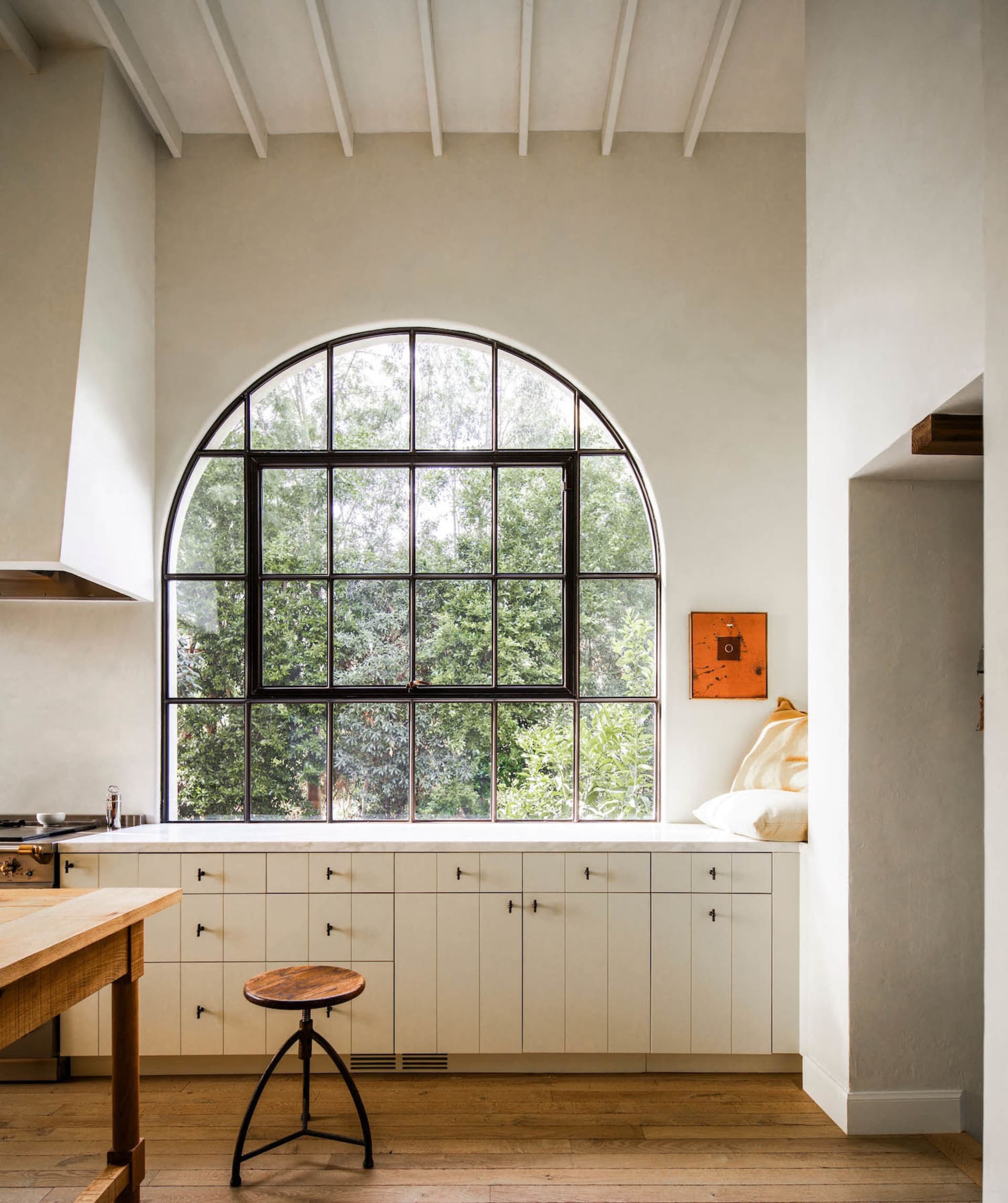A minimalist kitchen with white cabinets and a large arched window overlooking greenery.
