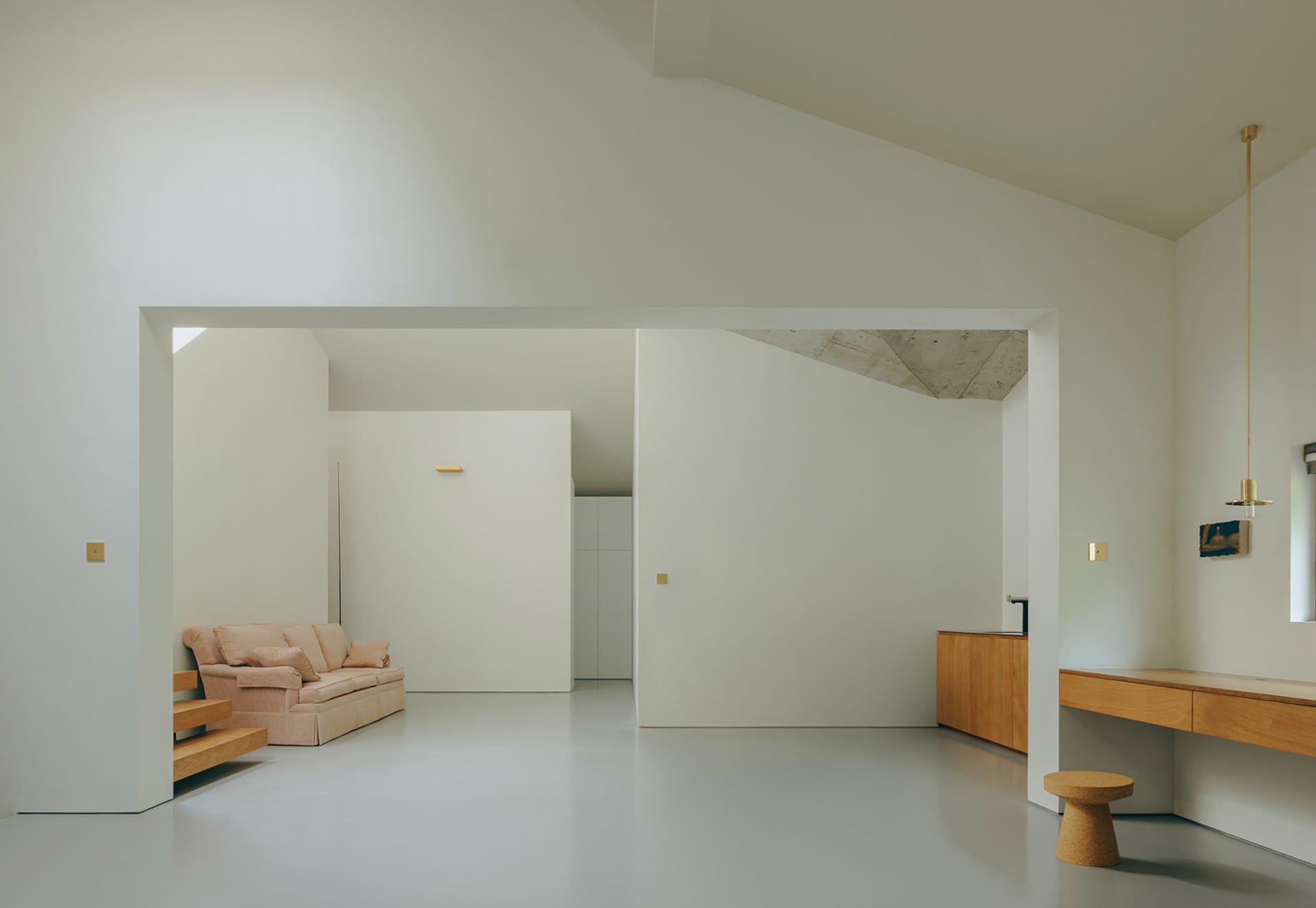 A minimalist living room with concrete and white walls, a low wooden bench, and a beige sofa with pillows.