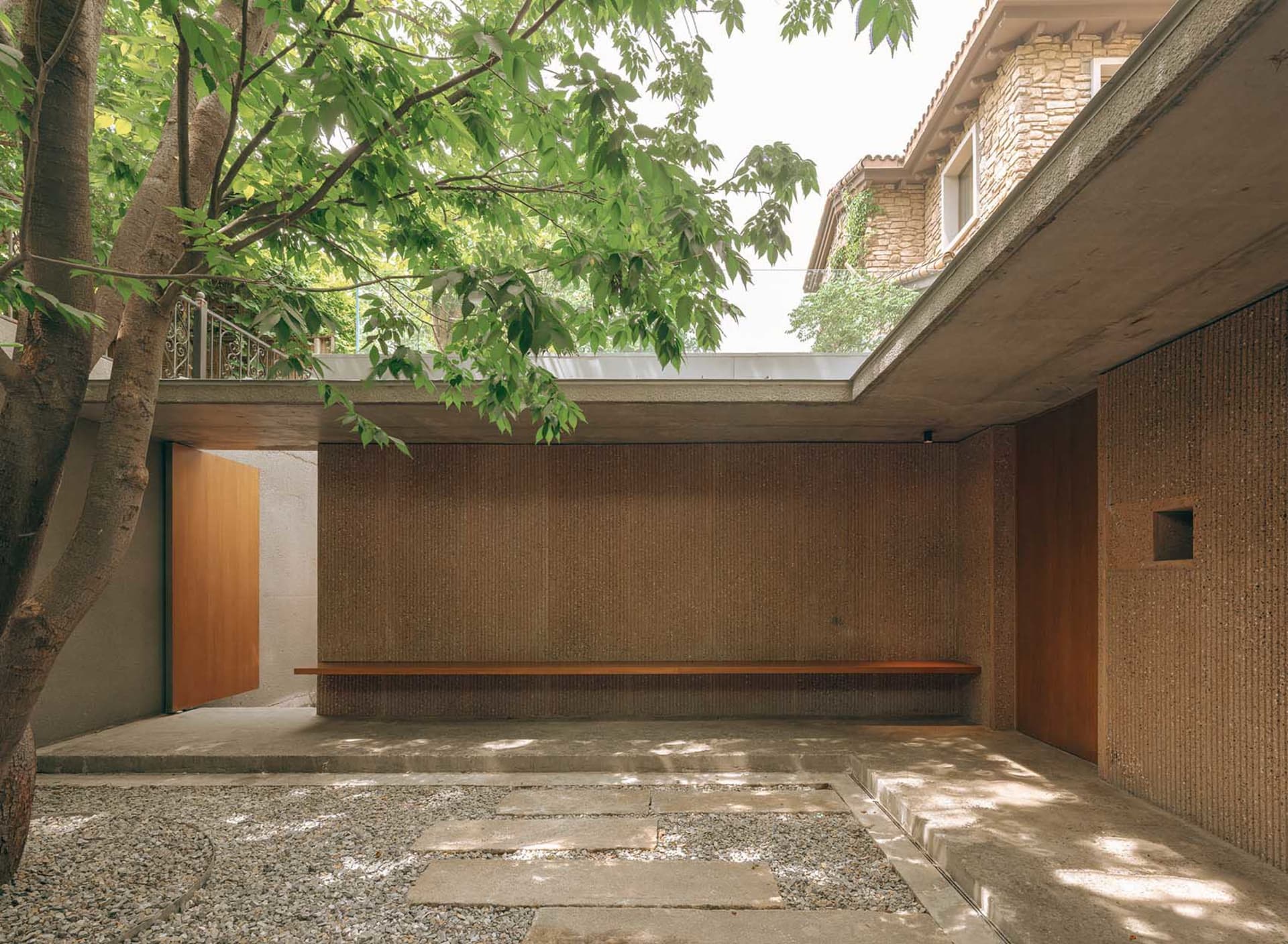 A minimalist residential patio featuring concrete flooring and wooden bench under a tree, with a roof providing shade.