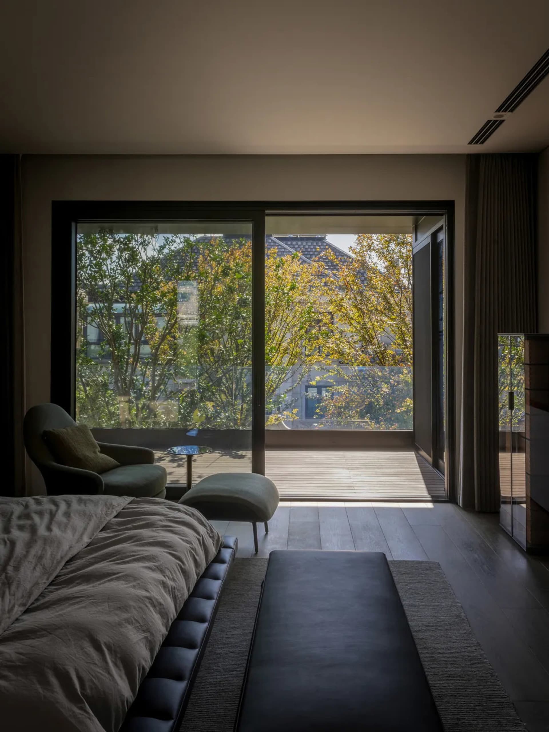 A modern bedroom featuring a large bed, a bench, and a large glass window overlooking trees and a deck.