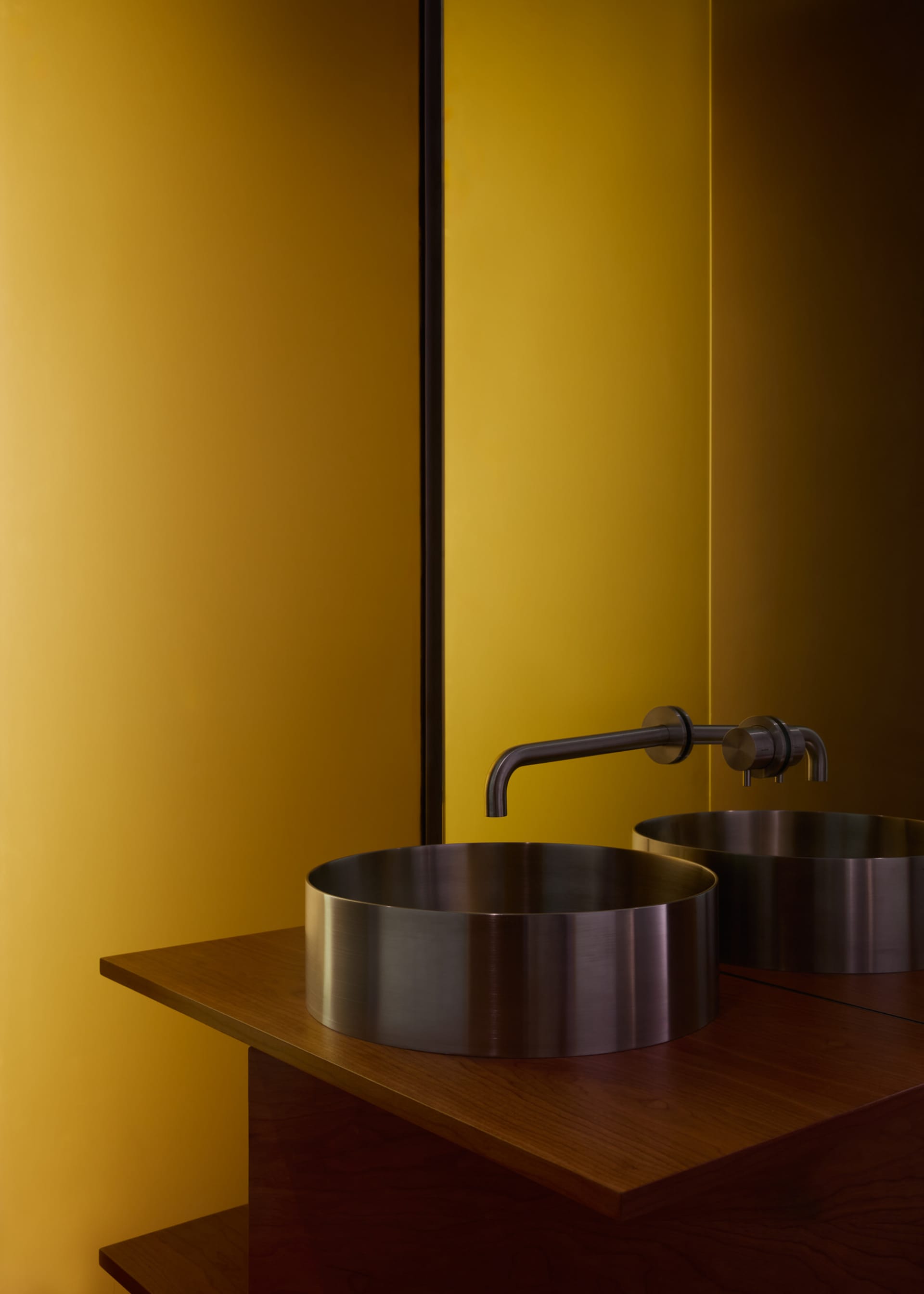 A modern metallic basin with a sleek, minimalist design against a vibrant yellow wall, featuring a single basin faucet.