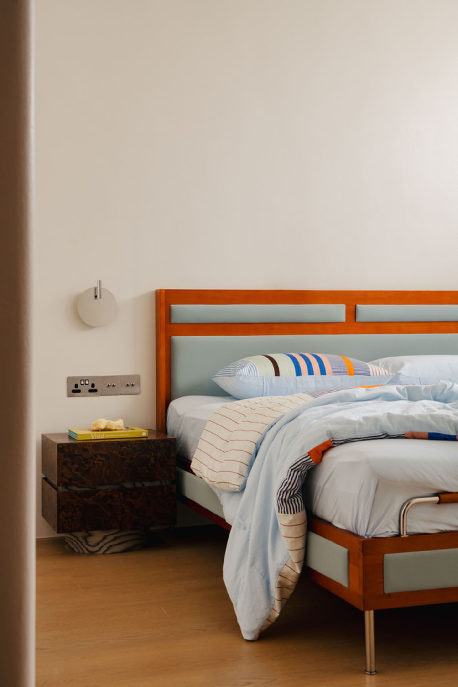 A minimalist bedroom featuring a wooden bedside cabinet and a bed with a light blue and white striped duvet, against a plain