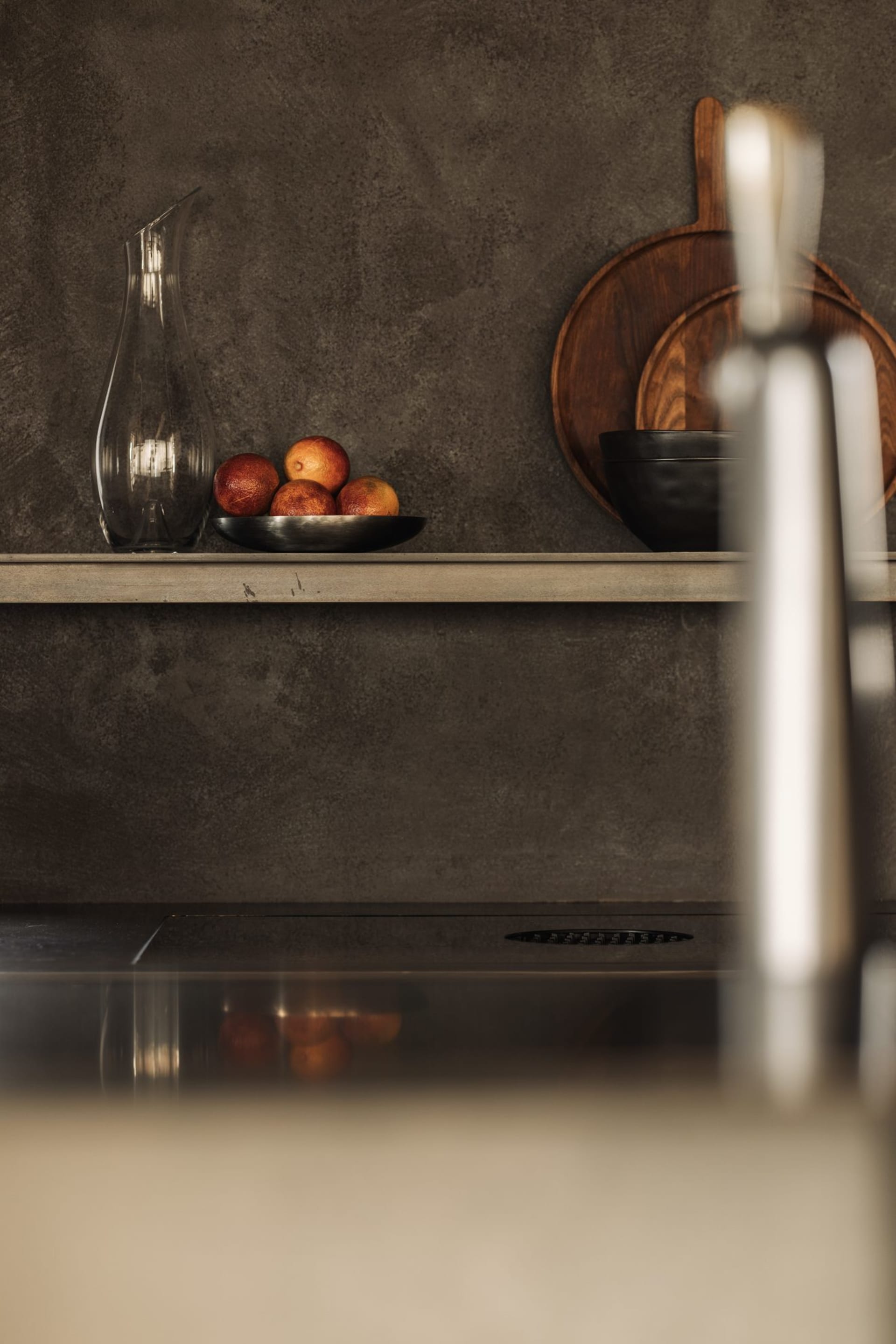A modern kitchen with a concrete countertop, featuring oranges and a pitcher on a floating shelf.
