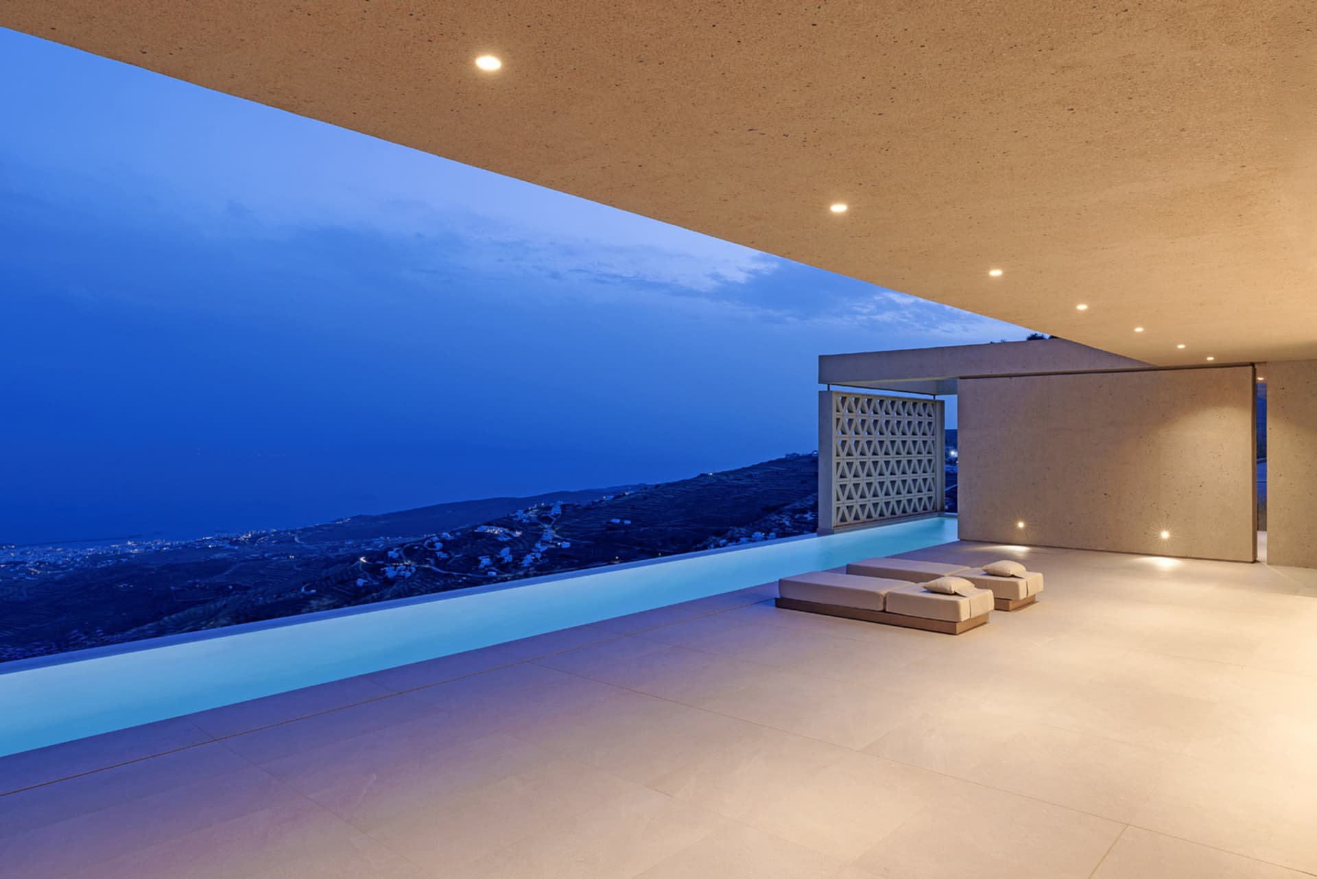 Modern pool deck with concrete walls and a rooftop infinity pool overlooking a night-time landscape with illuminated houses a