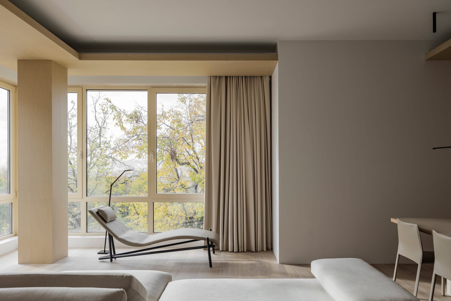 A minimalist living room with neutral fabrics and wood elements, featuring a lounge chair, sofa, and large windows with a vie
