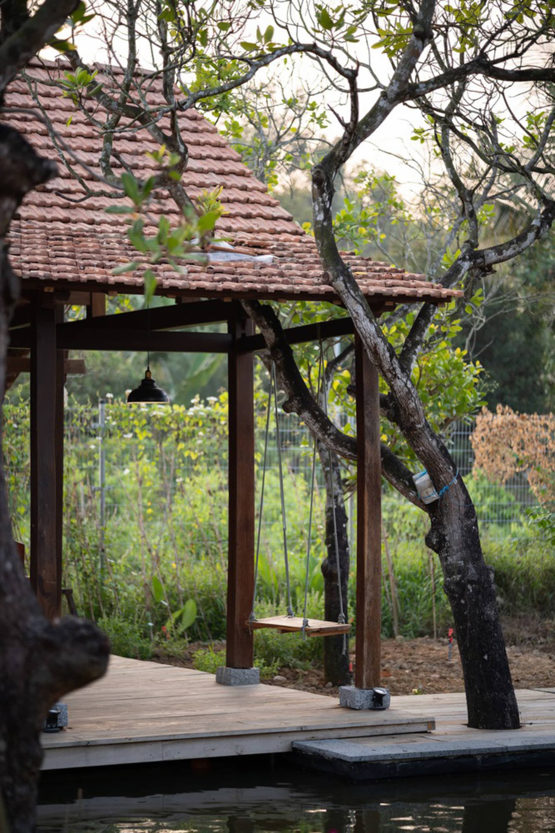 A wooden structure with a tiled roof and a hanging swing, surrounded by greenery and trees.