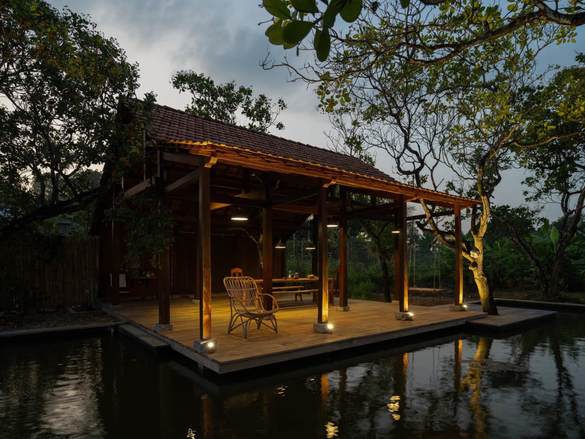 A wooden verandah with a thatched roof at dusk, floating over a pond, surrounded by trees and illuminated by warm lights.
