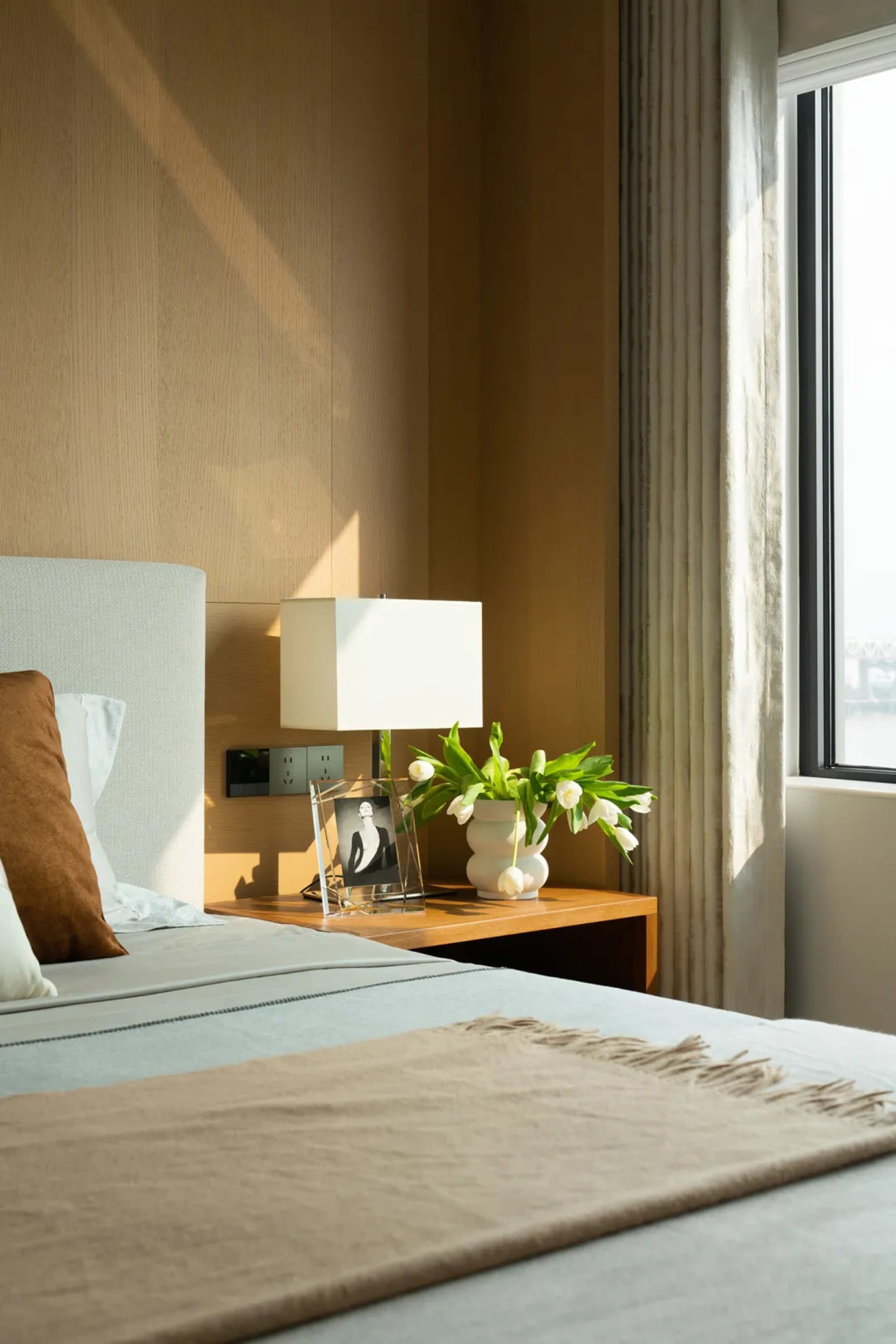 A modern minimalist bedroom featuring a wooden bed frame, fabric bedding, and a glass lamp and vase with white tulips.