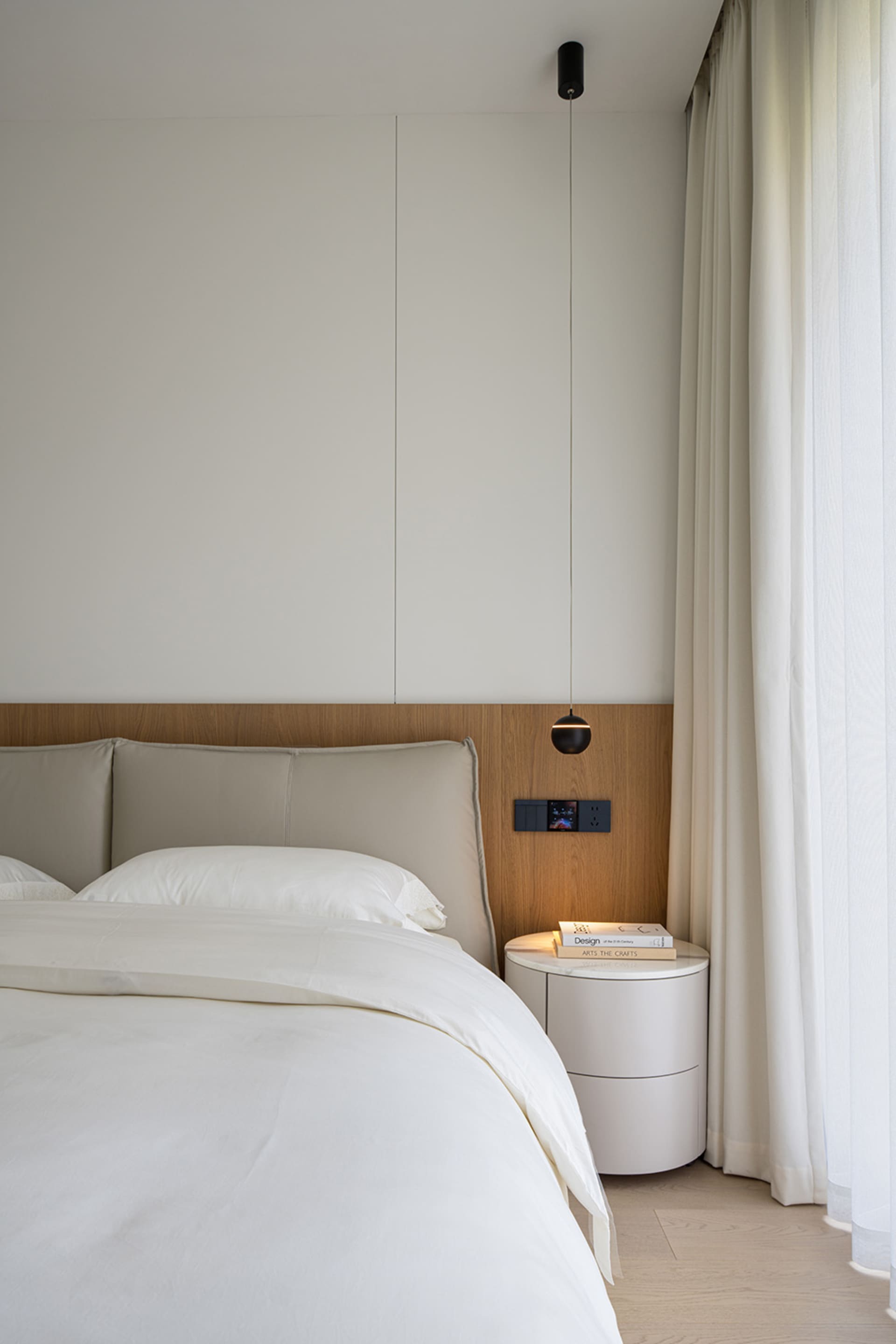 Modern minimalist bedroom featuring a white bed with grey headboard, wooden flooring, and sheer white curtains.