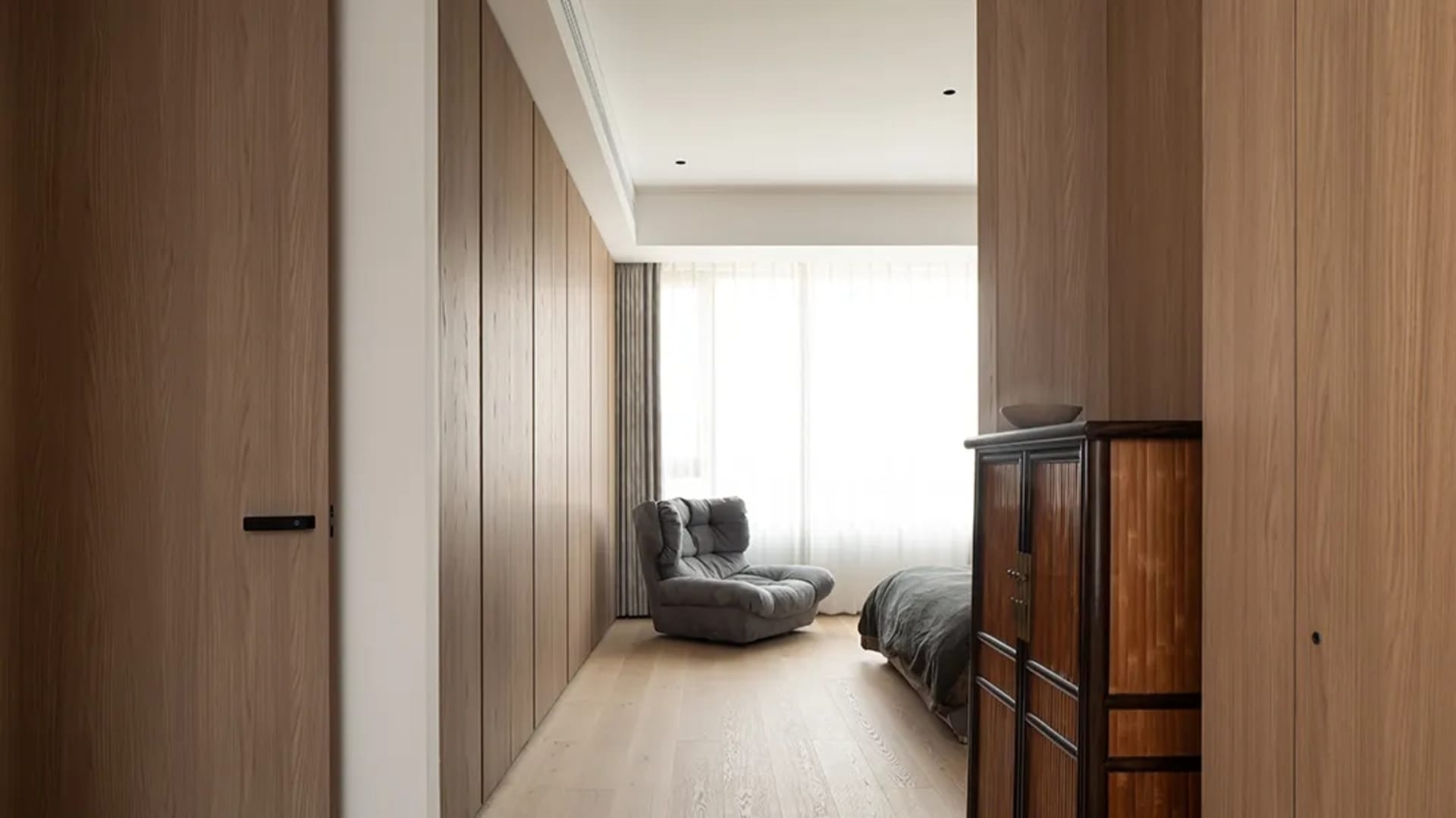 Minimalist bedroom featuring natural wood flooring and furniture, with a comfortable seating area and a window with sheer cur