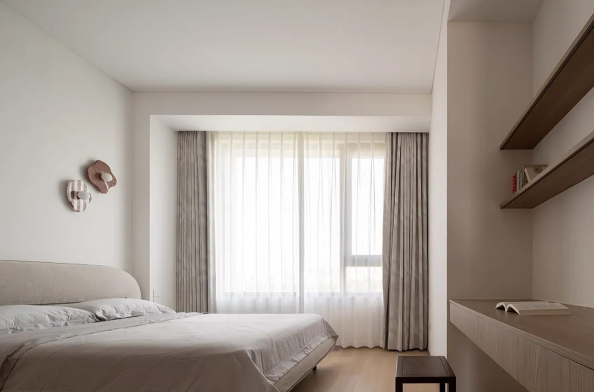 A modern bedroom with fabric headboard, wooden sides of the bed and floating desk on the wall, featuring sheer curtains over