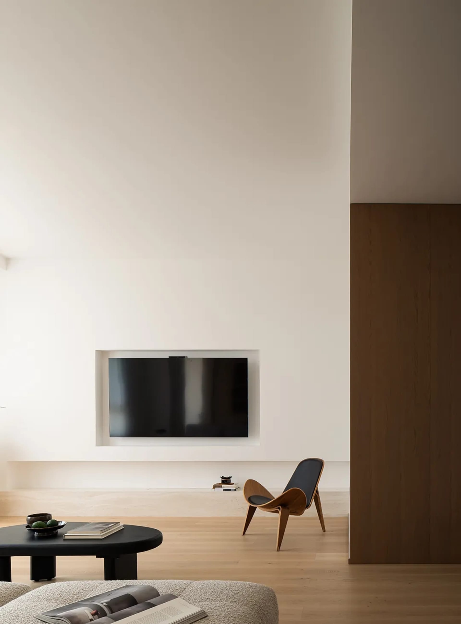 Modern minimalist living room with a sleek black television embedded in the wall, a wooden shell chair, and a black ottoman w
