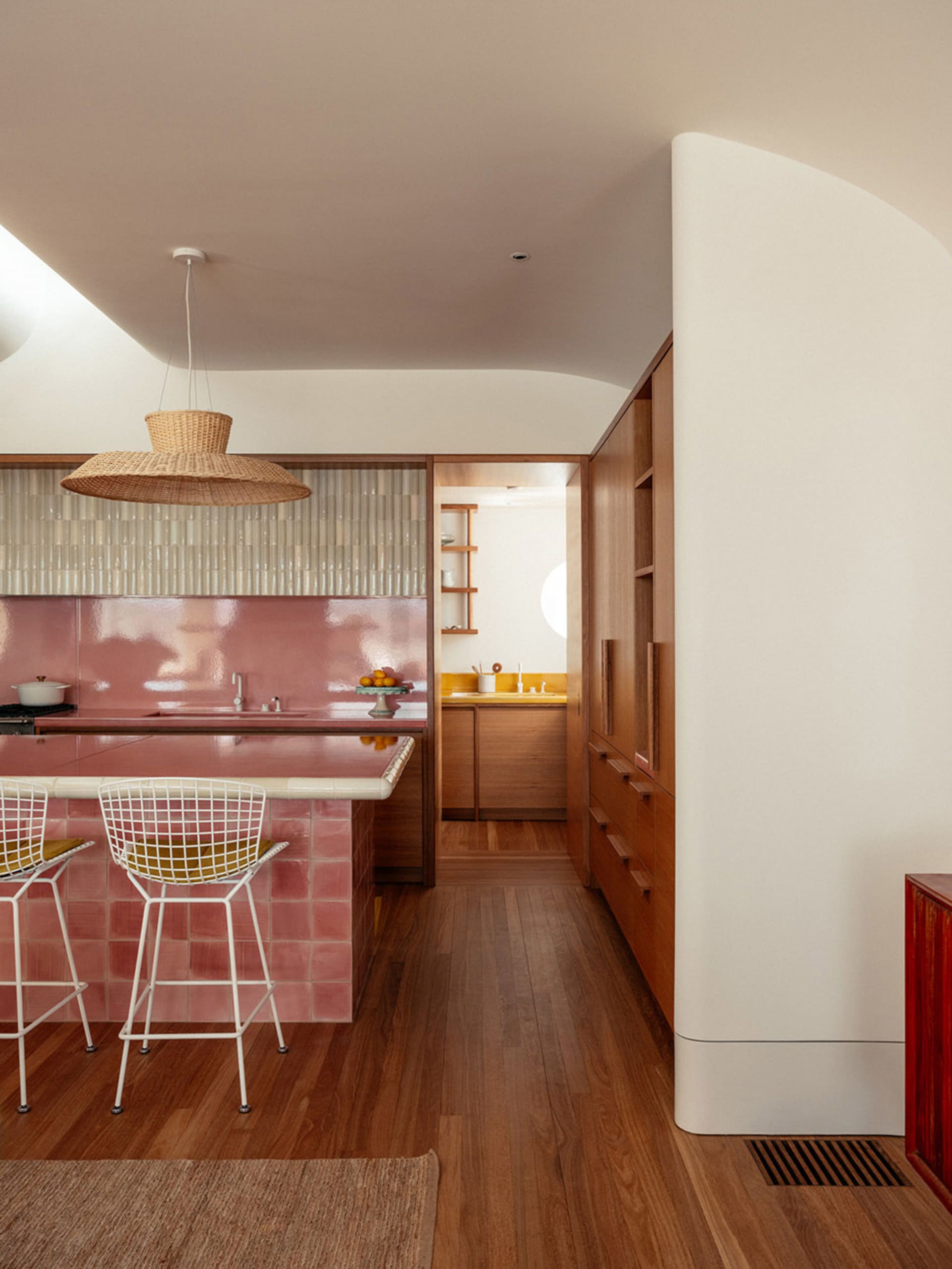 A modern kitchen with wooden floors, pink tiles, and white bar stools.