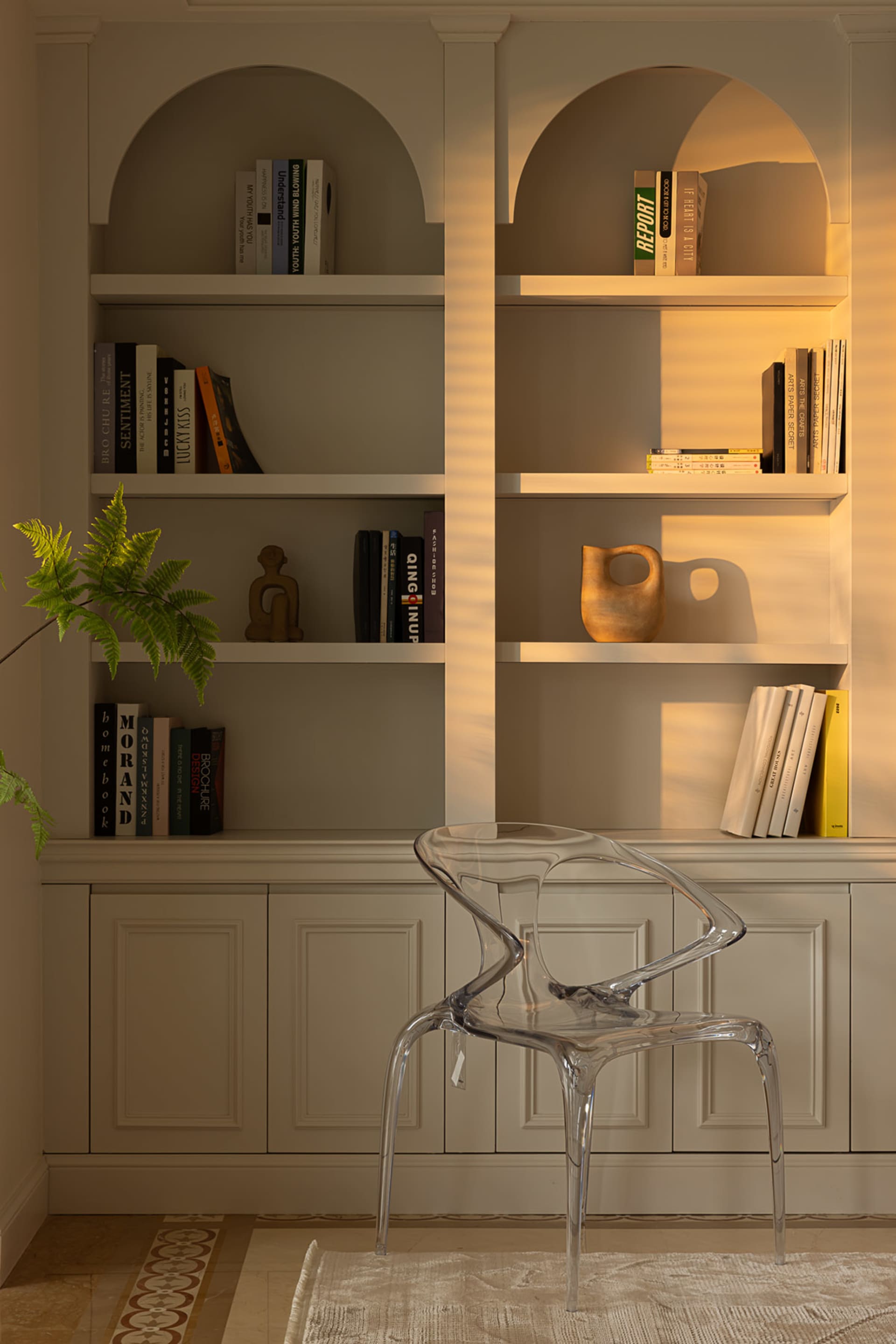 A minimalist library with a glass chair, white shelves, wooden vase, and white painted walls and panels.