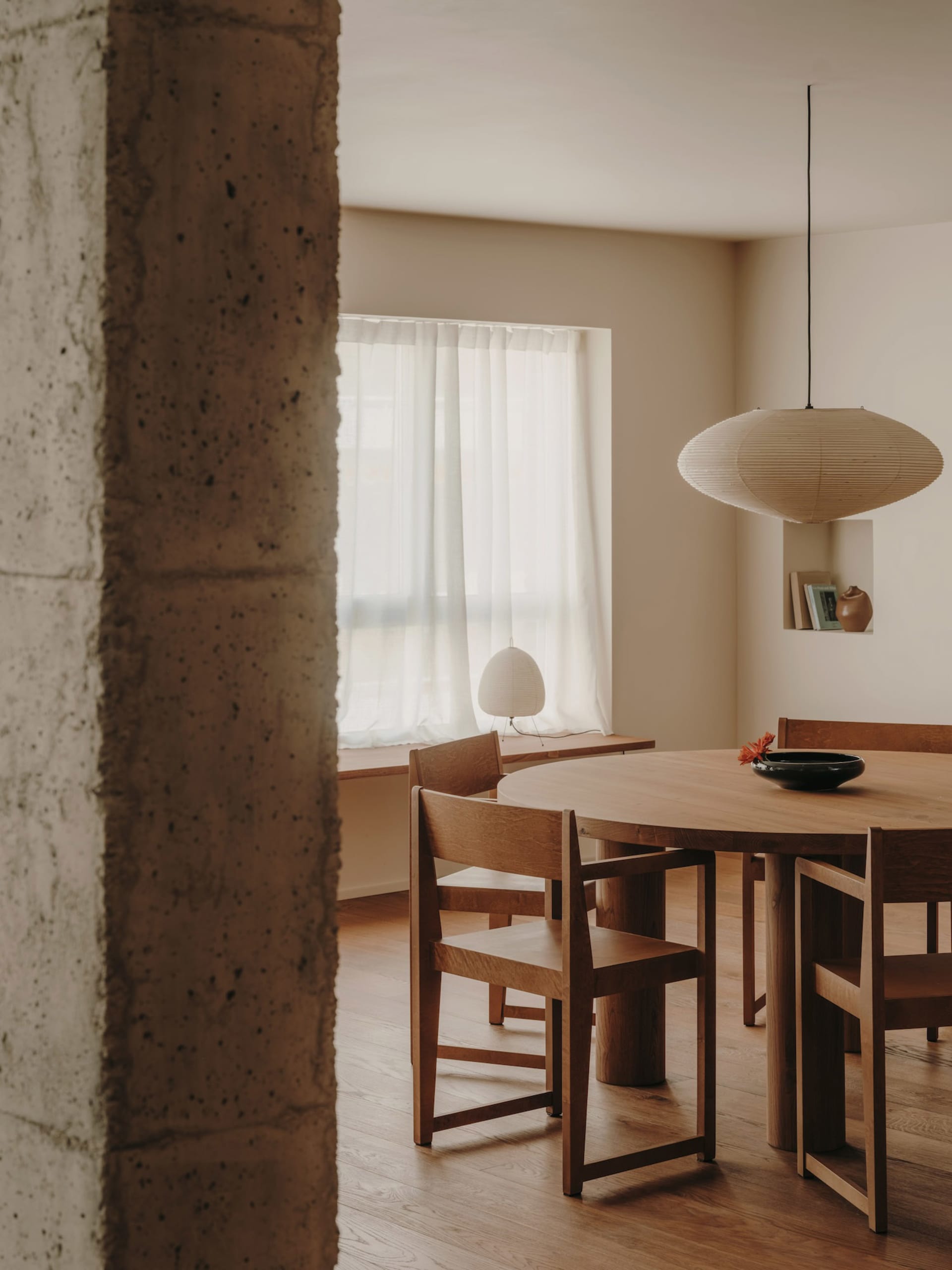 A minimalist kitchen features a wooden table and chairs, a large window with sheer curtains, and a modern pendant lamp.