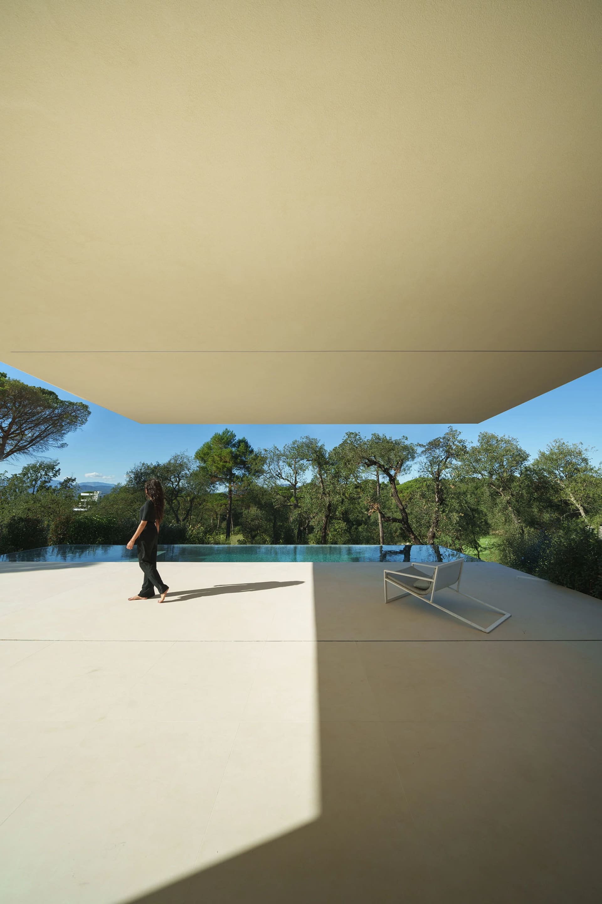 A modern minimalist terrace with concrete flooring and a large infinity pool surrounded by lush greenery.