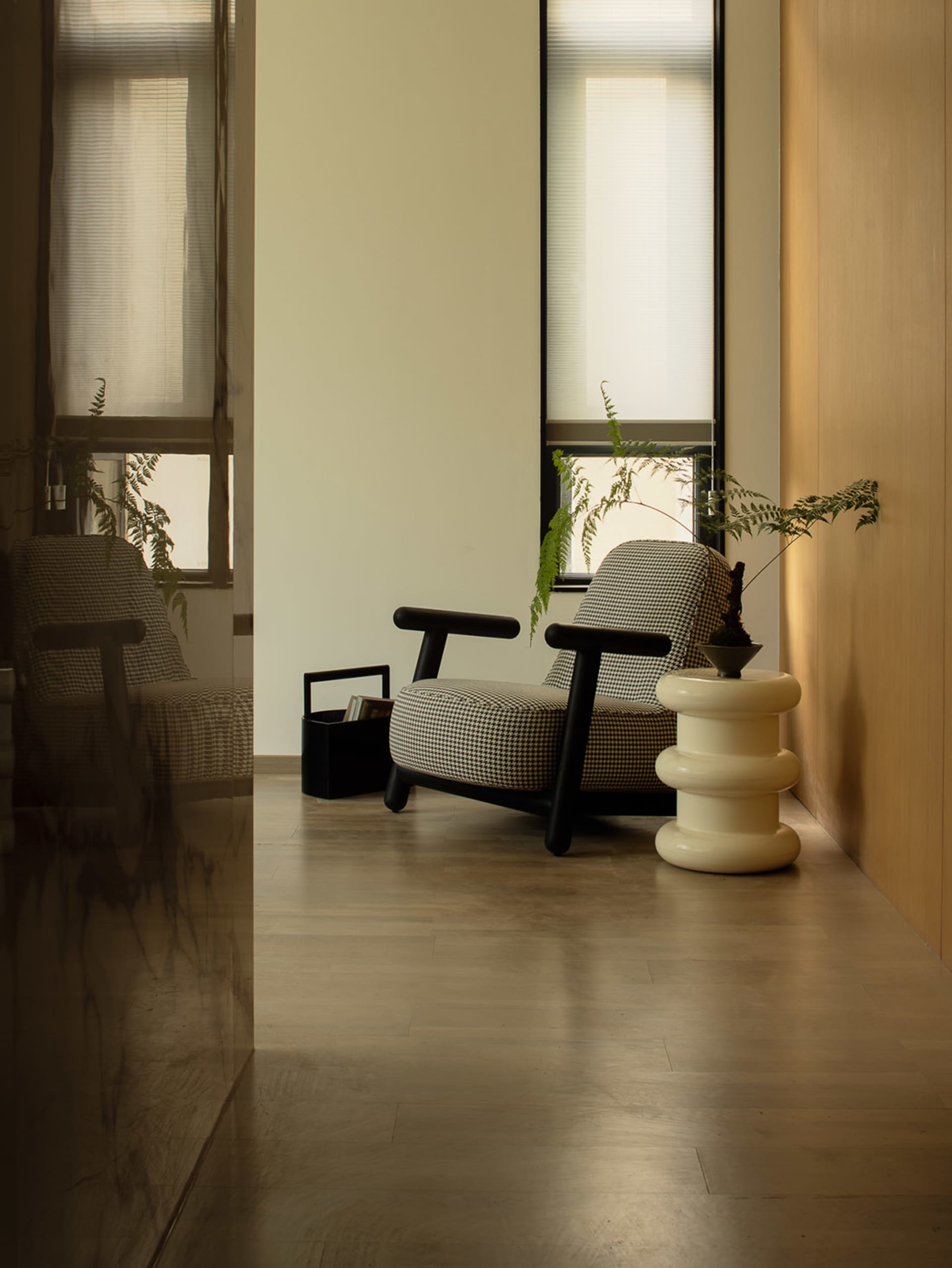 A minimalist monochrome living room with wooden floors and white walls, featuring a checkered armchair, side table, and plant