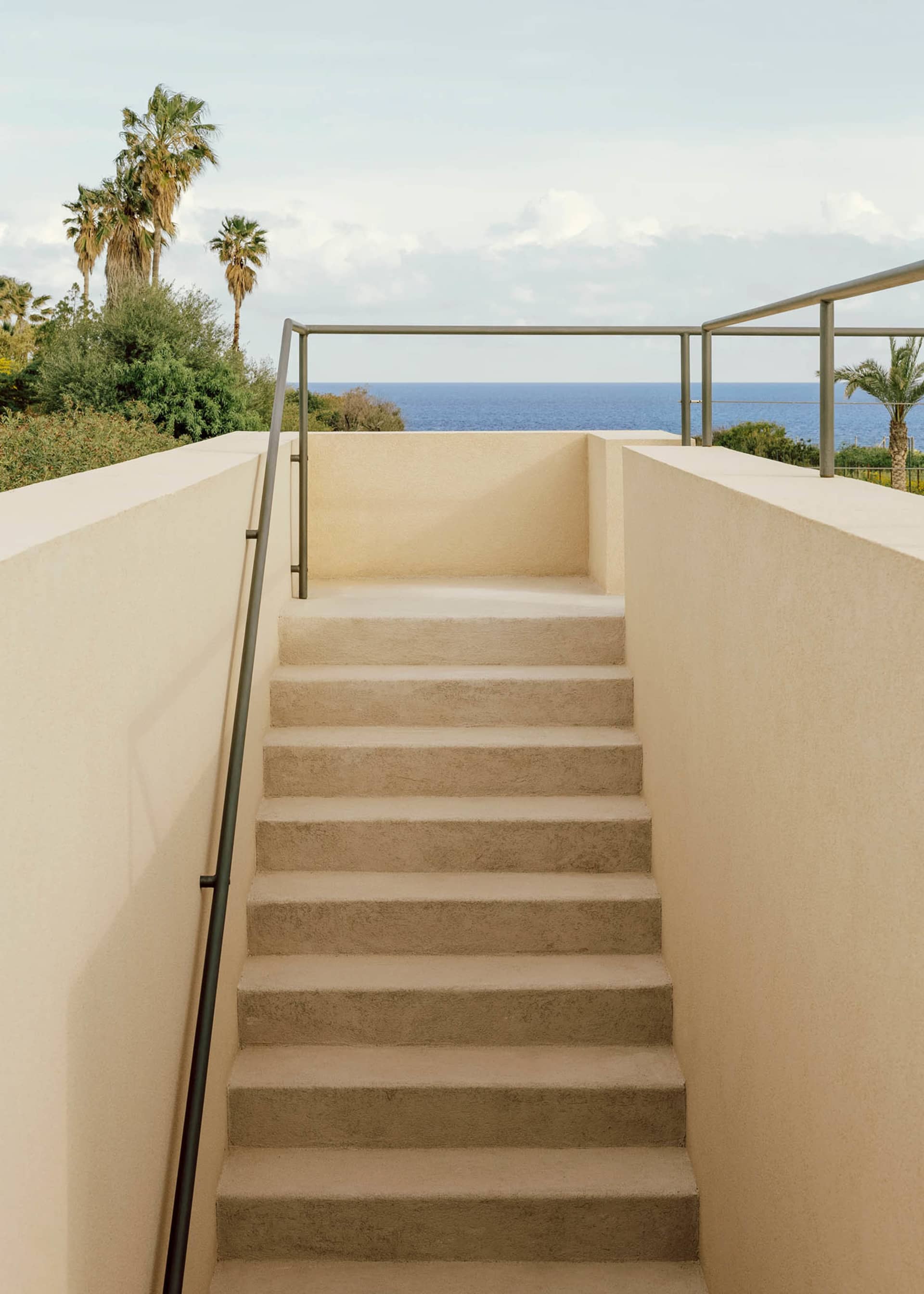 An exterior staircase with concrete steps and railings overlooking the ocean with palm trees in the background.