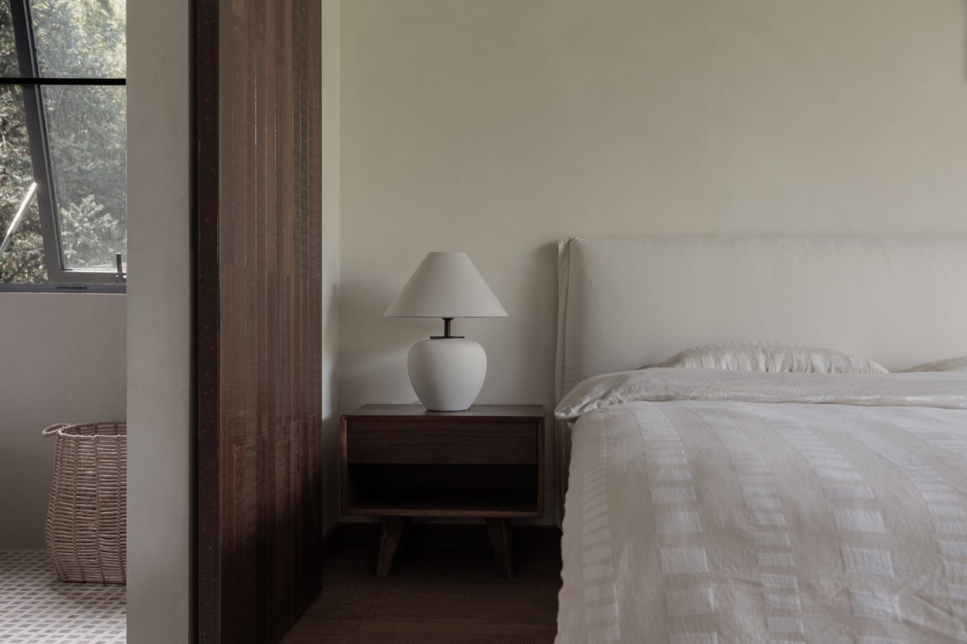 A modern minimalist bedroom with a white fabric bedspread and a minimalist bedside table with a white shade lamp.