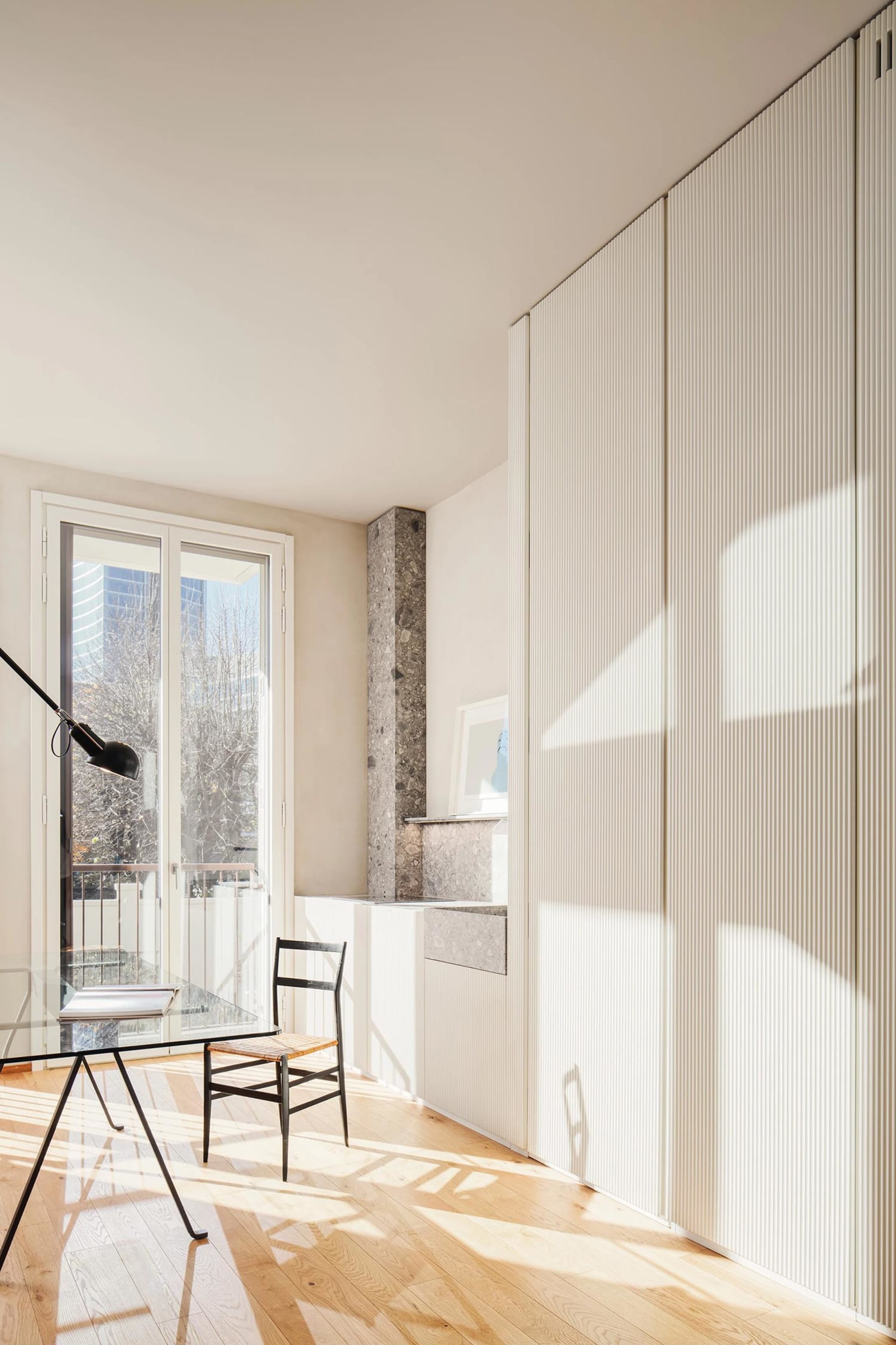 A modern home office featuring wooden flooring and a glass table with a black lamp and a wooden chair.