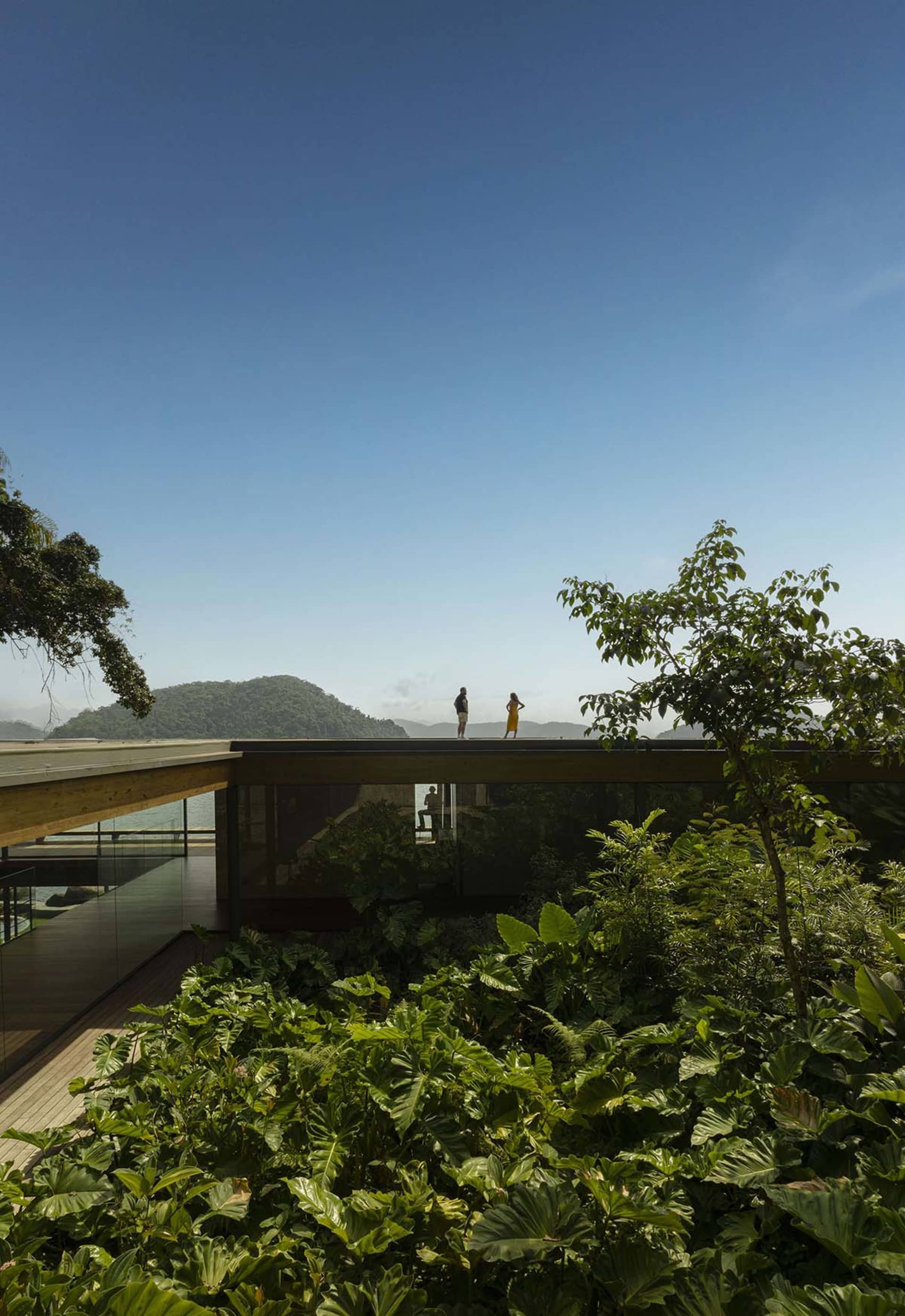People standing on a modern veranda overlooking lush greenery and distant mountains under a clear blue sky.