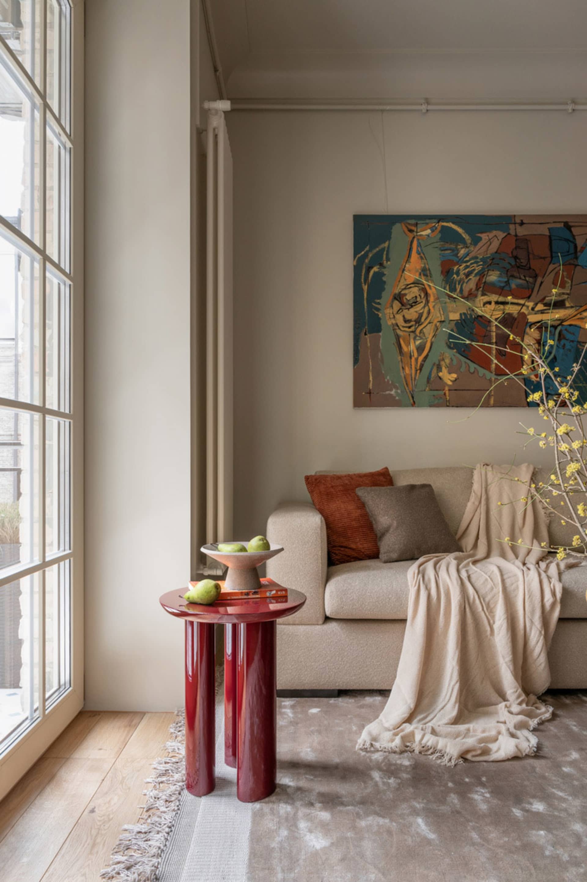 A modern living room featuring a contemporary art piece on the wall, a beige couch with cushions, and a red cylindrical table