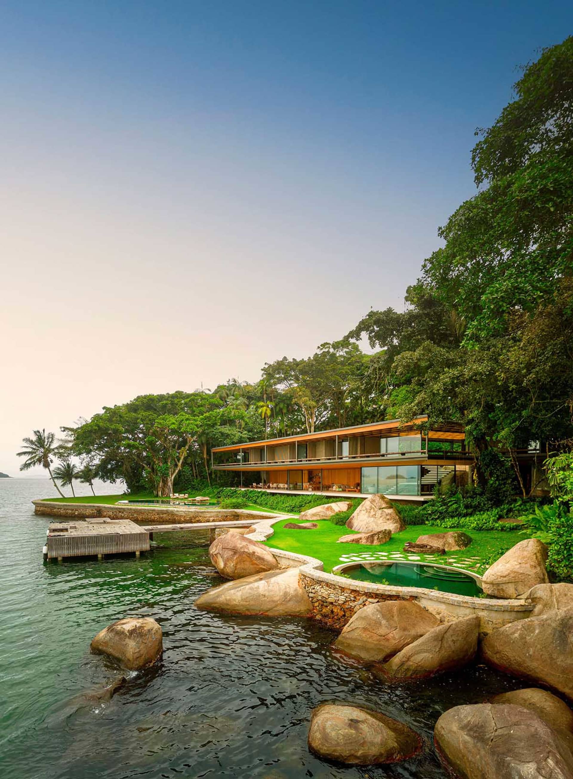 Modern residential home situated on the water, featuring a contemporary design with wooden exterior, glass walls, and stone a