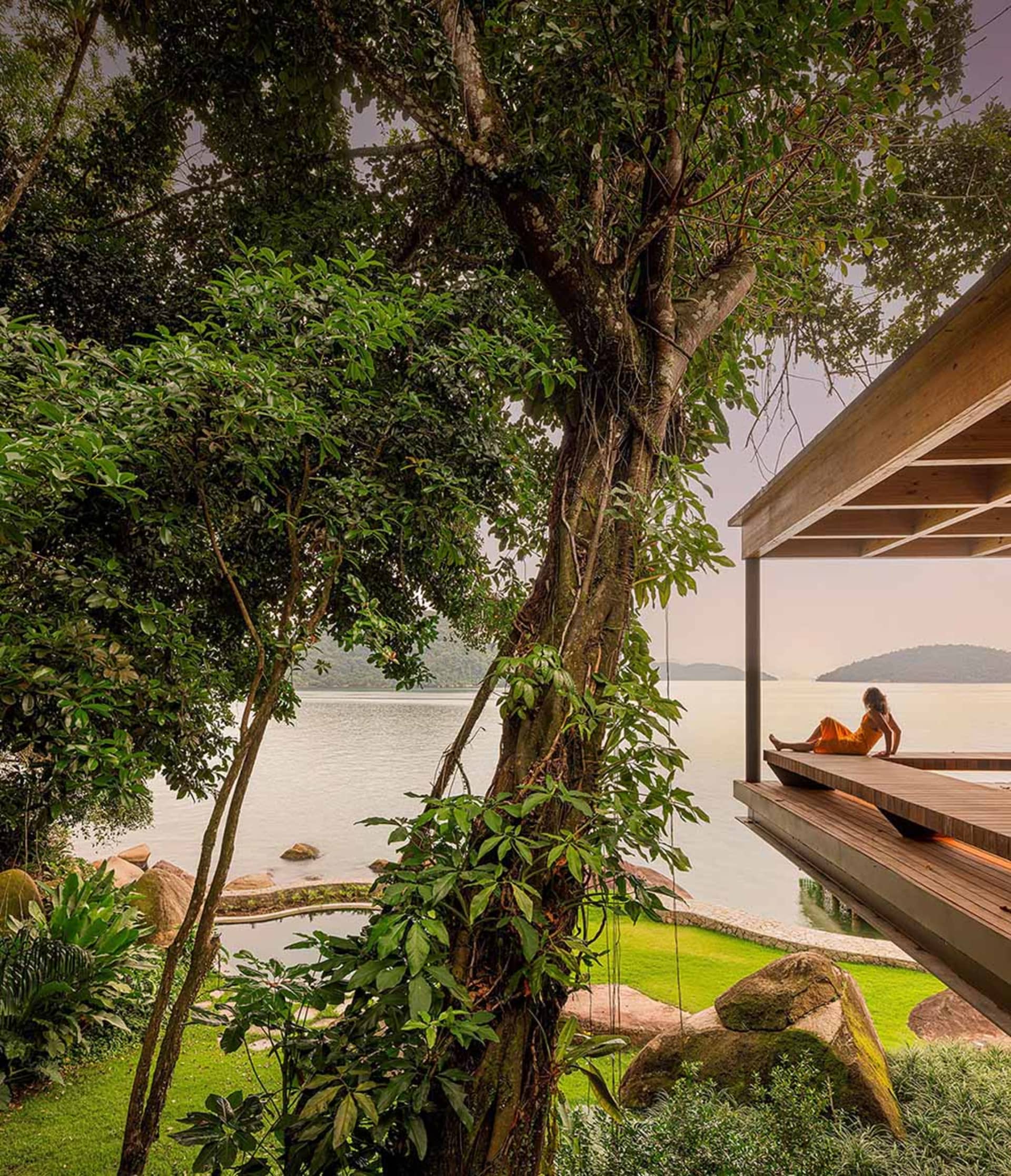 A person in an orange dress sitting on a wooden deck overlooking a serene lake surrounded by lush greenery and rocks.