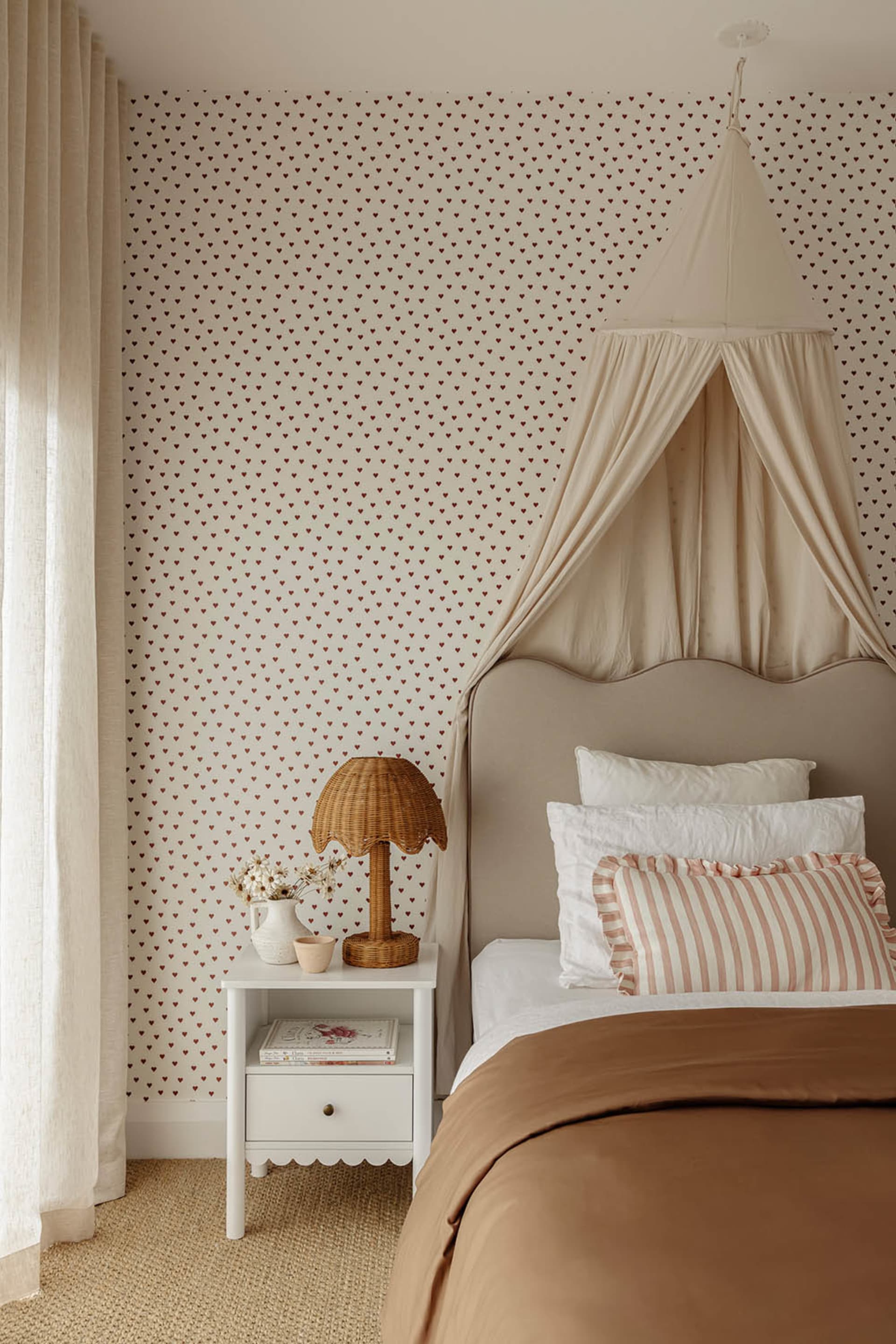 A cozy bedroom featuring a canopy bed with soft fabrics, a side table with a wicker lamp and white pitcher, and heart-pattern