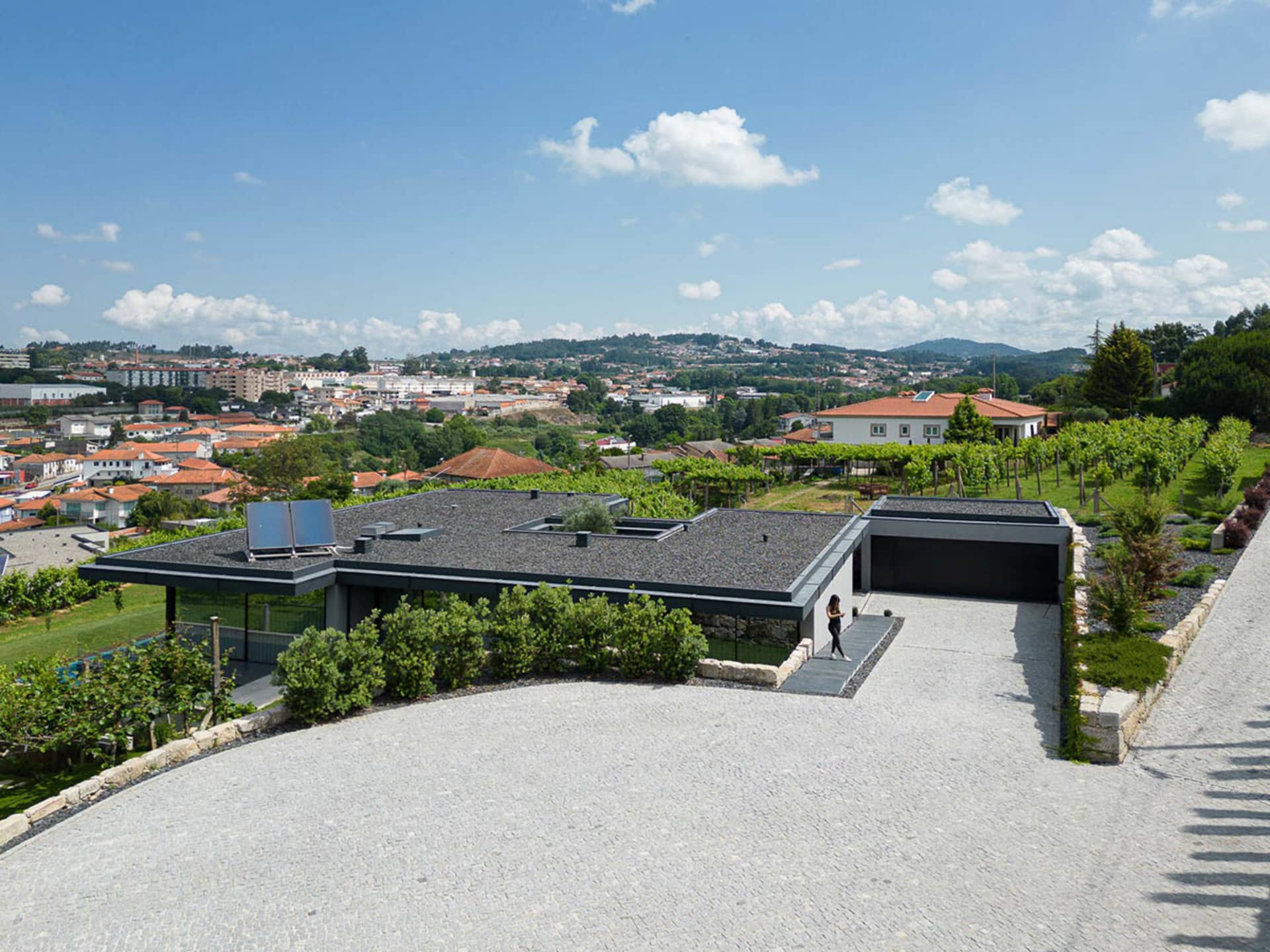 A modern villa with concrete exterior and a sunlit driveway overlooking a town.