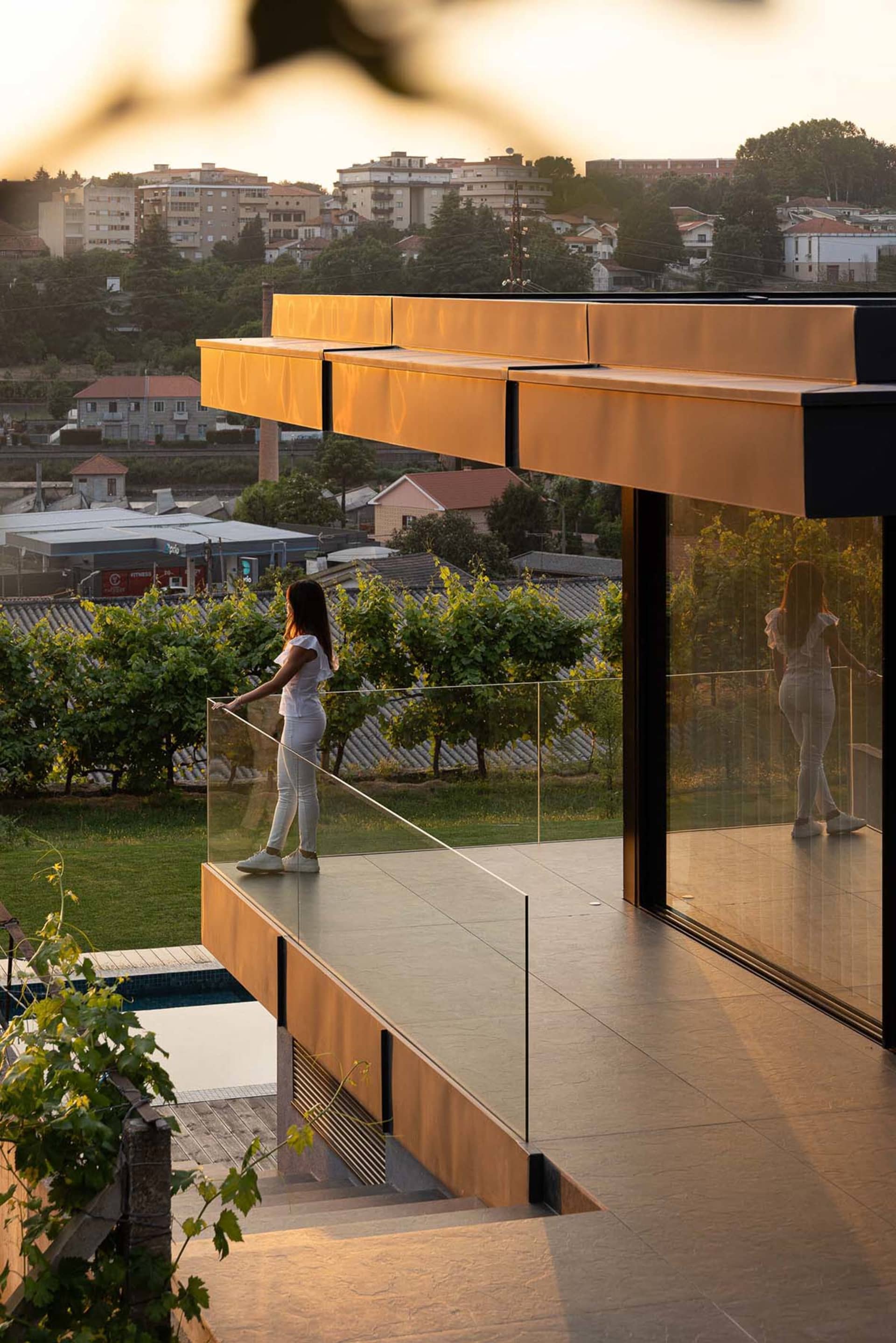 A woman stands on a modern veranda with glass panels and concrete flooring, overlooking a cityscape at sunset.