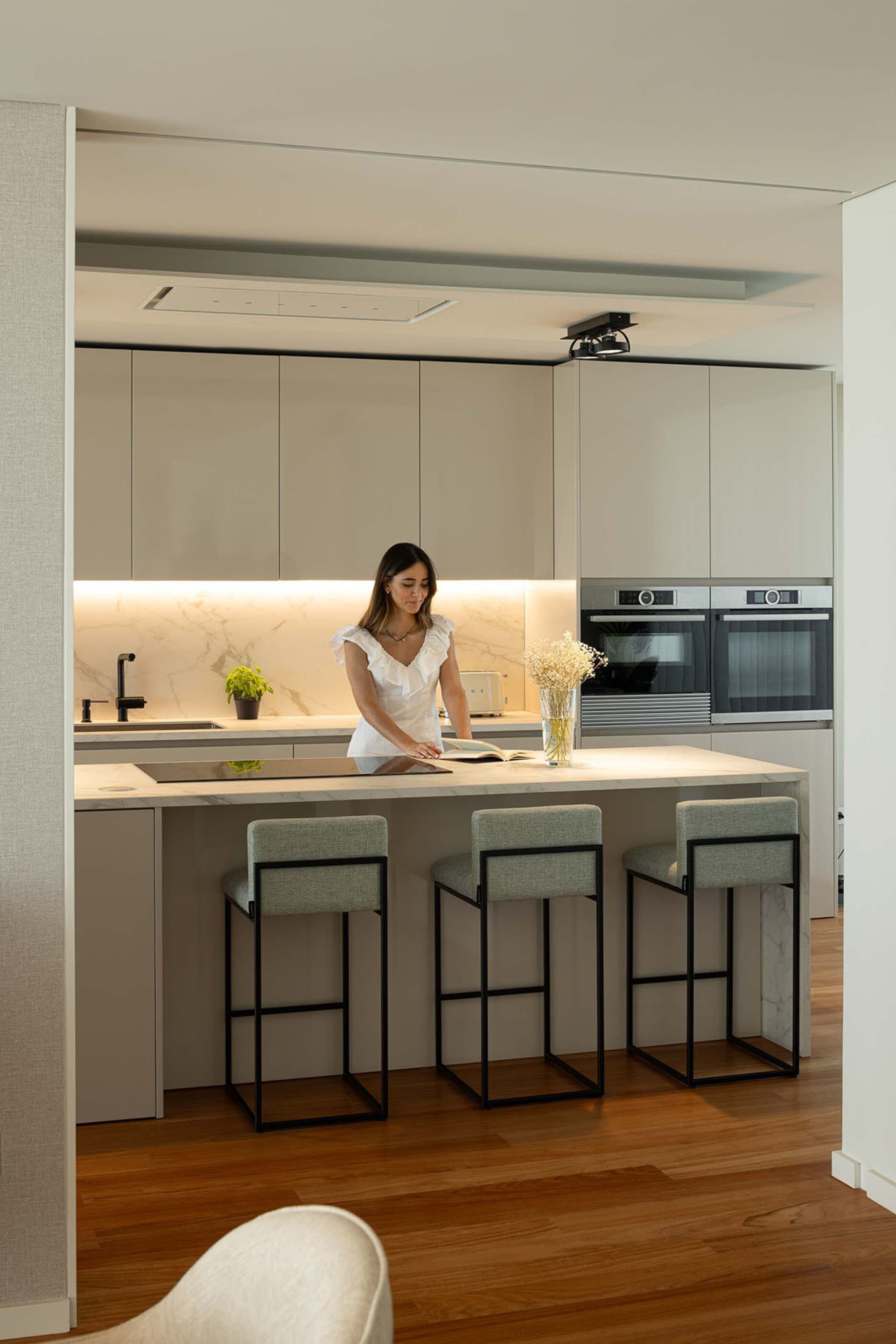 A modern minimalist kitchen with light-colored marble countertops, stainless steel appliances, and upholstered bar stools.