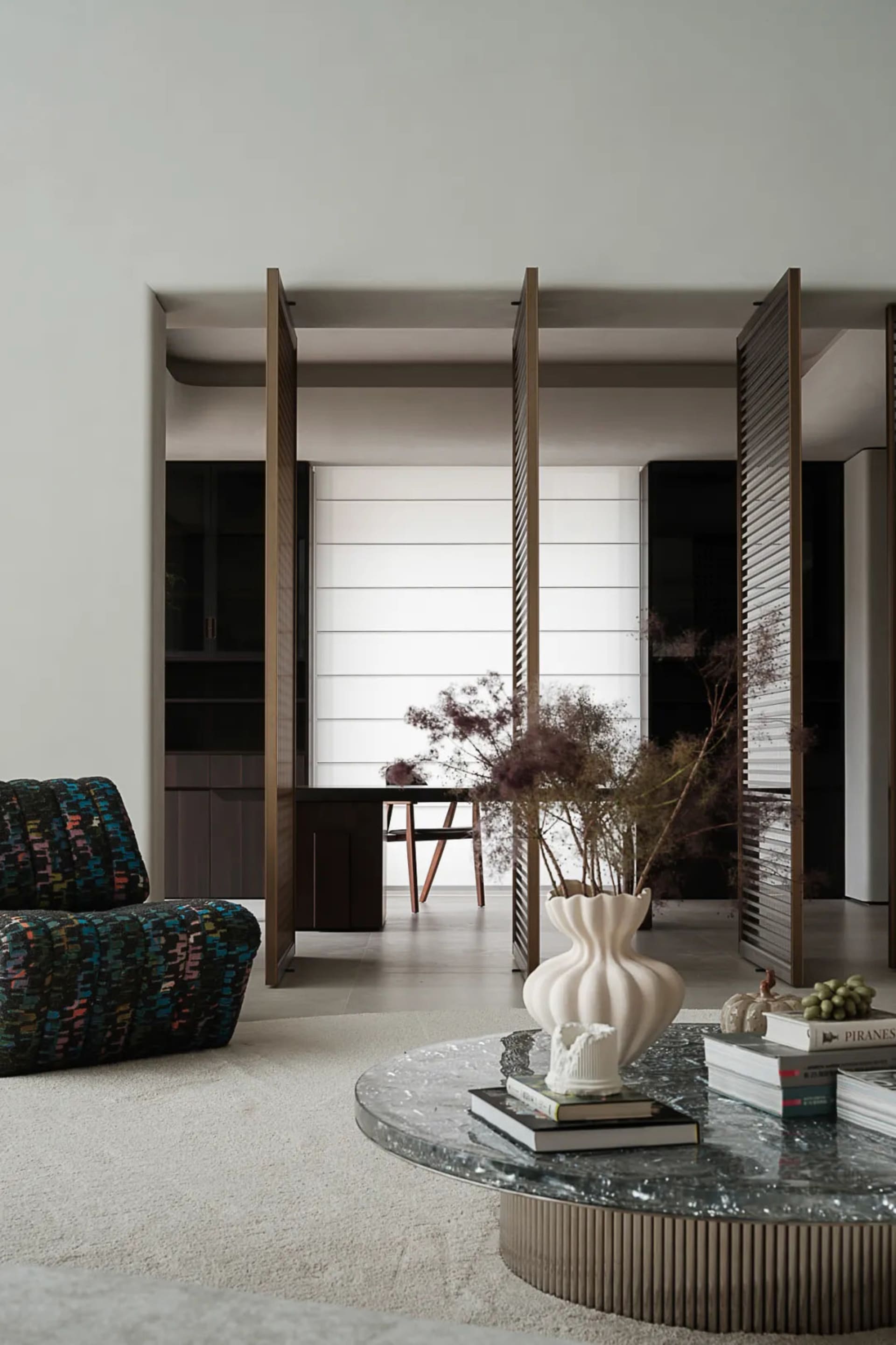 A modern minimalist living room with a round marble coffee table, a colorful patterned chair, and a white vase with dried pla