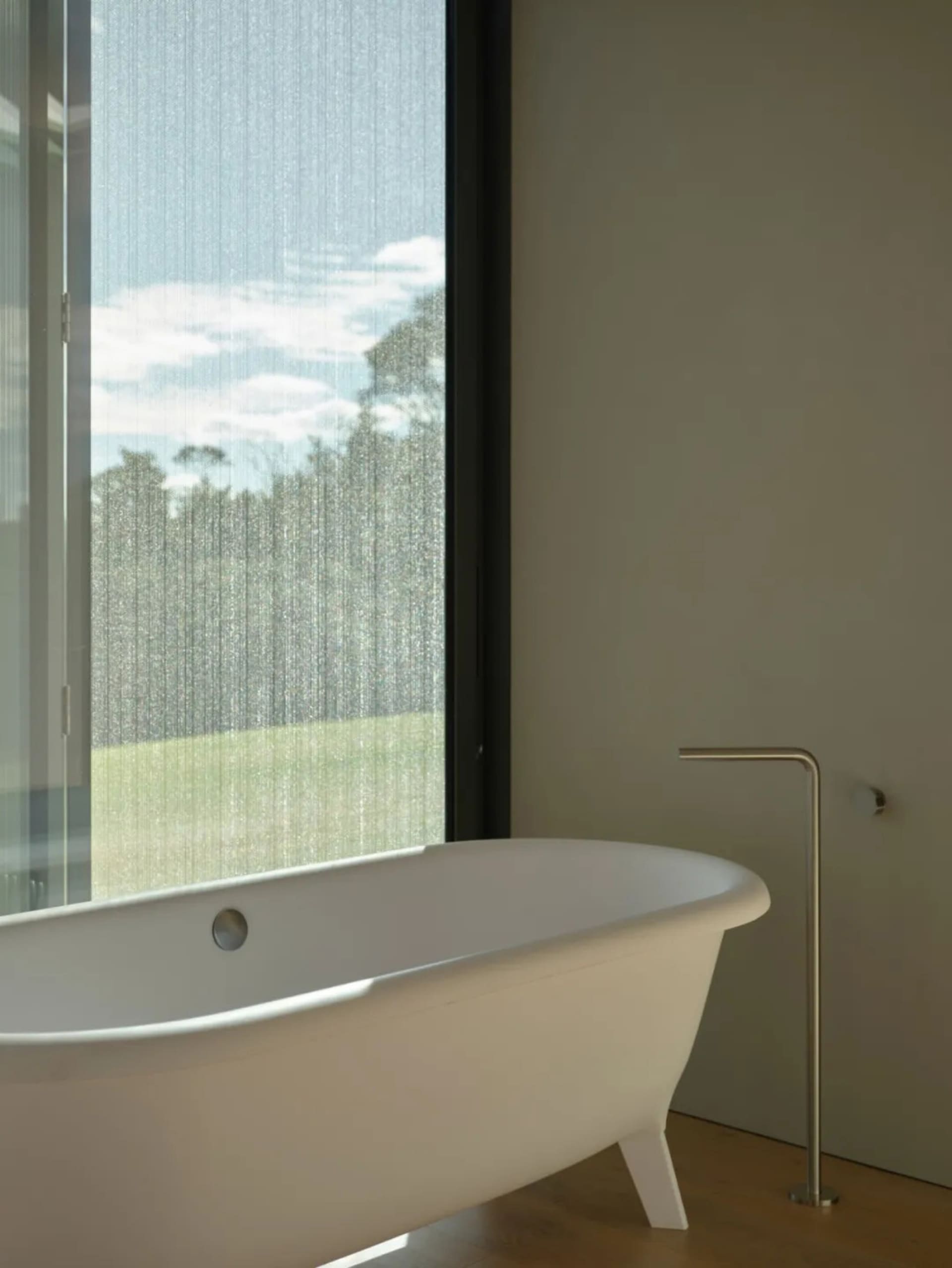 A minimalist white bathtub with a freestanding faucet in a bathroom with a large window showcasing a green landscape.