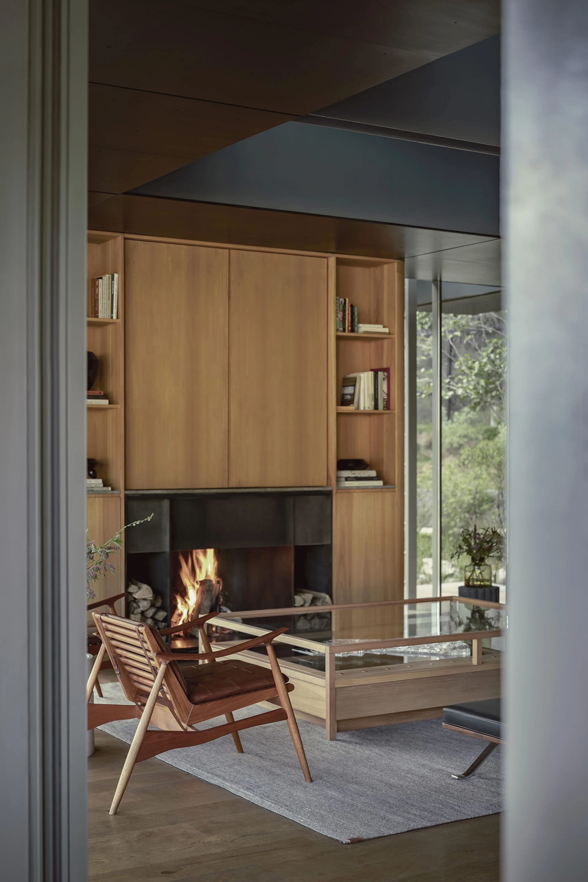 A modern living room with a wooden interior design featuring a fireplace, bookshelves, and a glass coffee table.