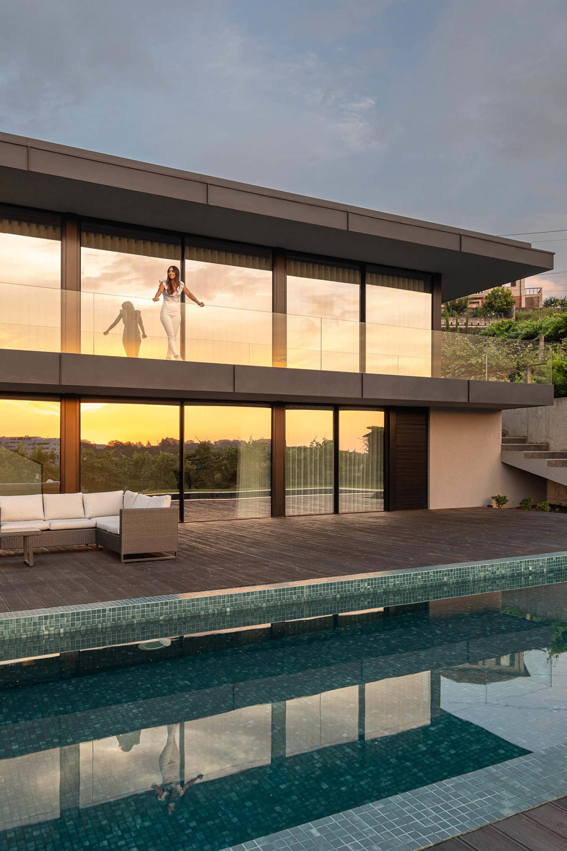 Modern residential architecture featuring glass walls and wooden deck with a swimming pool and sunset view.