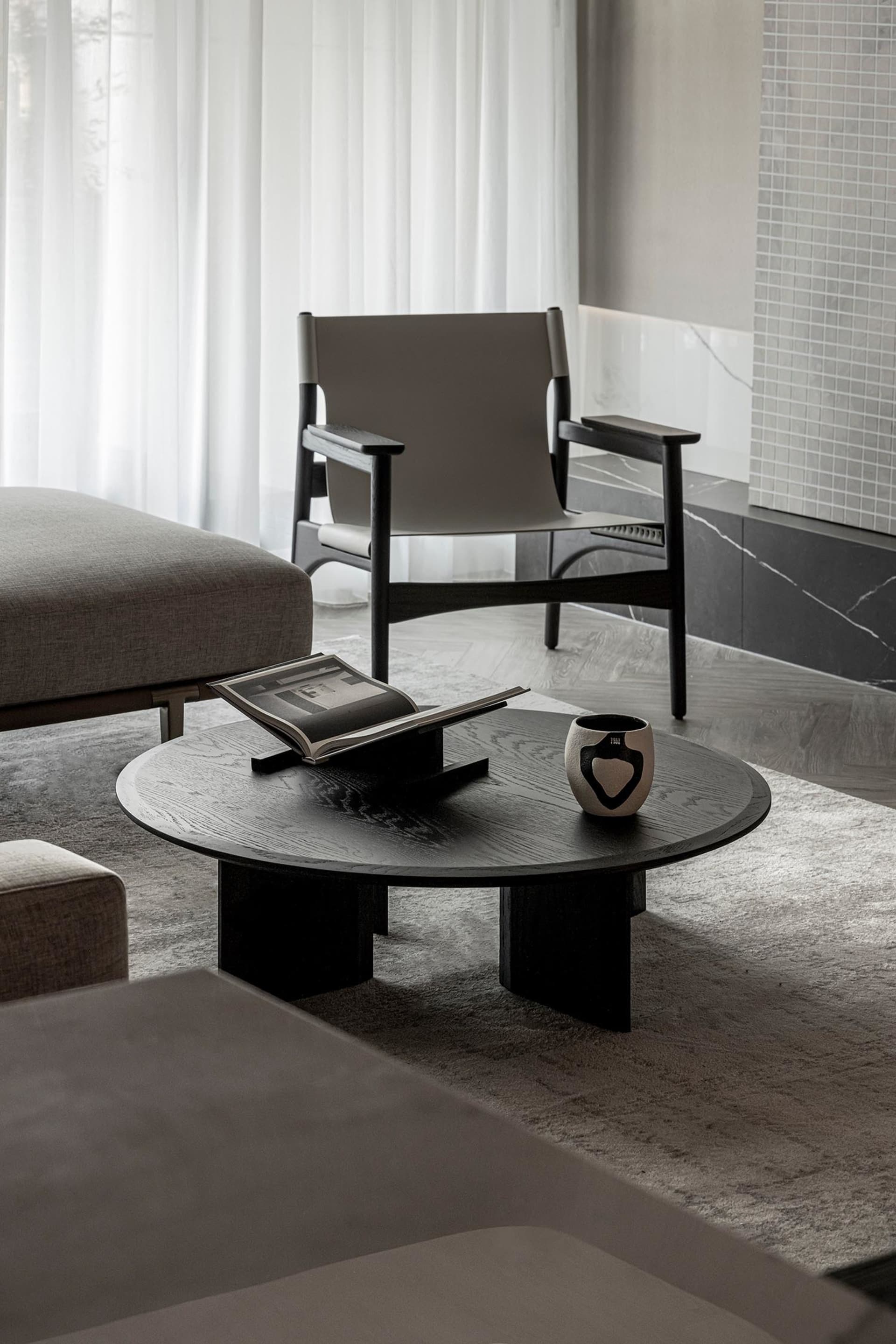 A minimalist living room scene with a round black coffee table, an open book, and a ceramic vase. A grey upholstered armchair
