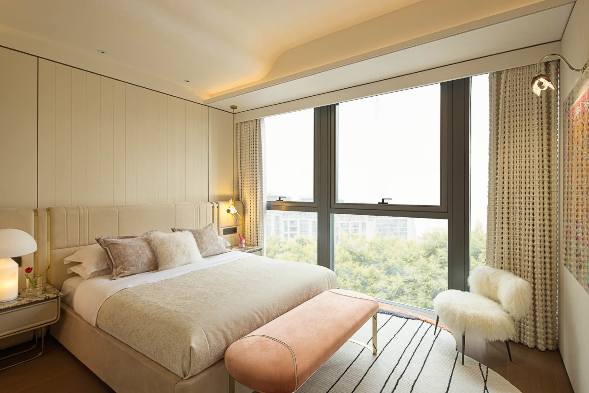A modern bedroom with a large window, a comfortable bed with plush bedding, and a cozy seating area with a fur-covered chair