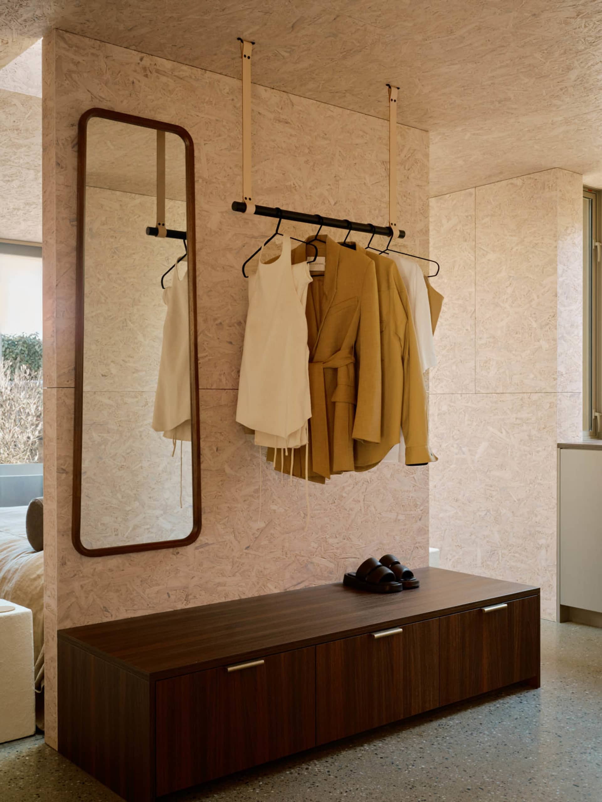 A modern closet with a wooden bench, jacket hanger, and mirror against textured wooden panels.