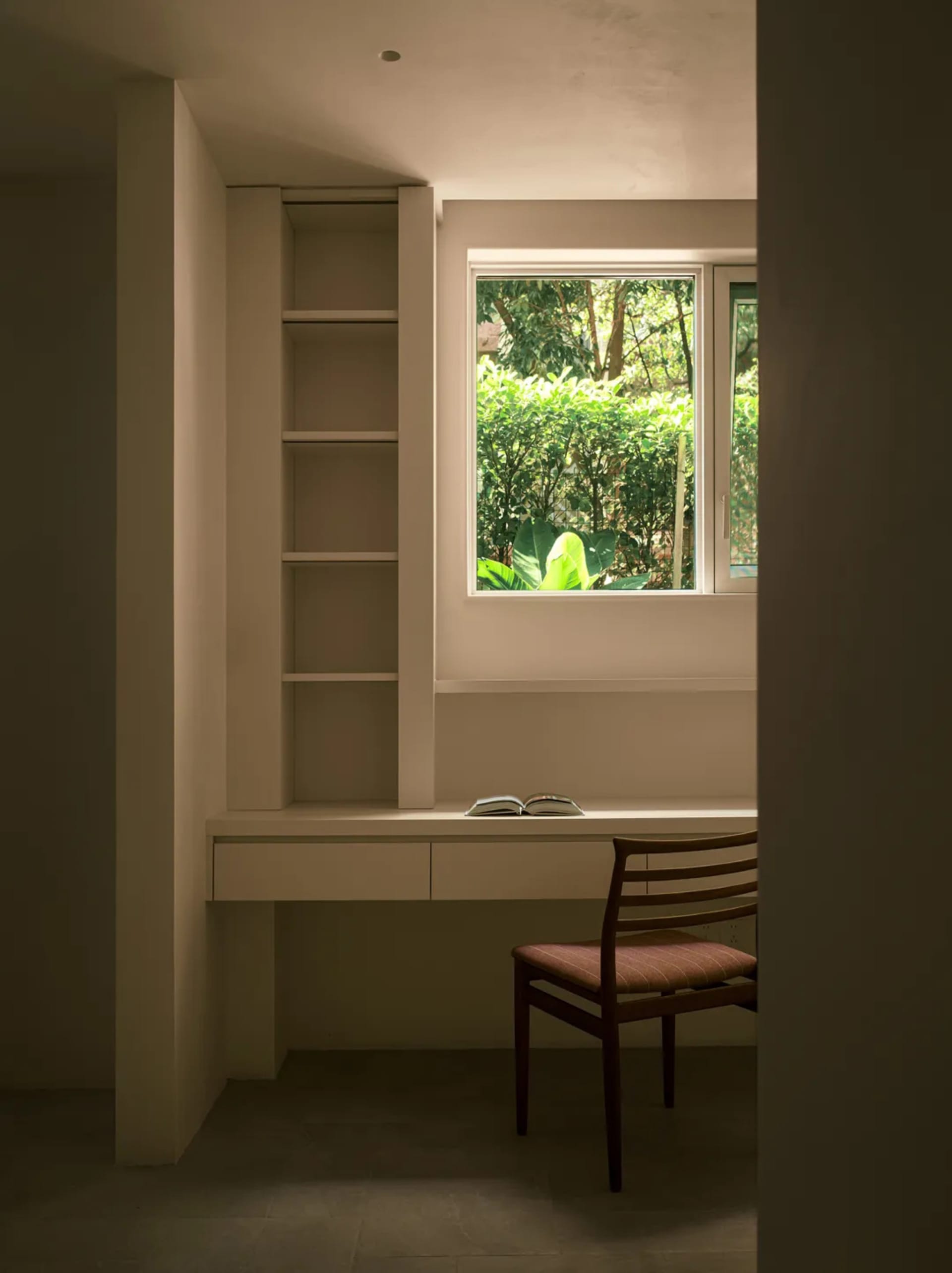 A white study room with a built-in desk, shelves, an open book on the desk, and a mid-century chair.