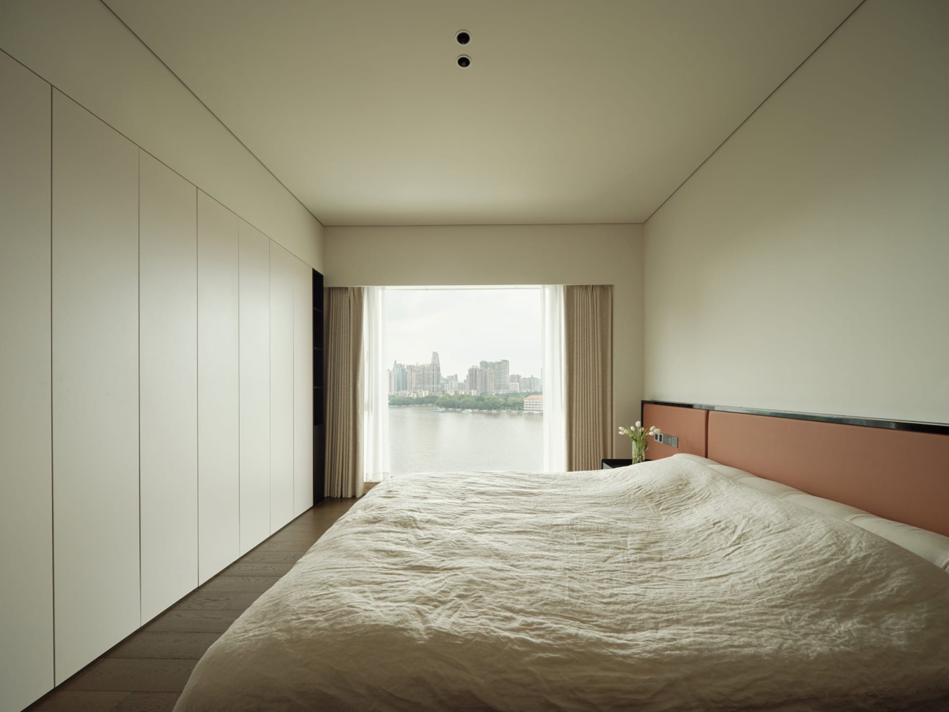 A minimalistic bedroom featuring fabric bedding and wooden accents with a view of the cityscape through the window.