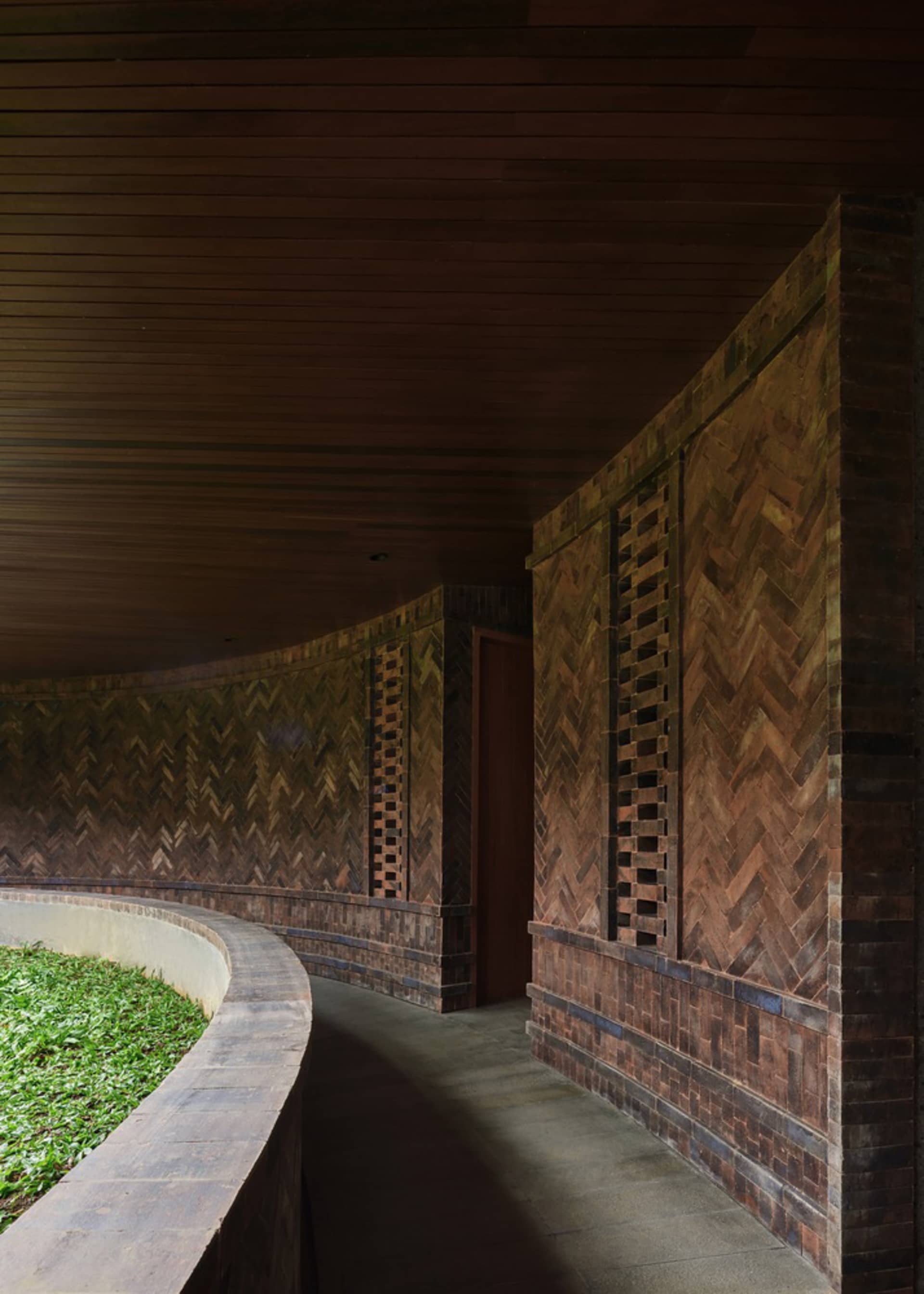 A curved corridor with wooden walls and brick walls, featuring a dark wooden ceiling and greenery along the bottom edge.