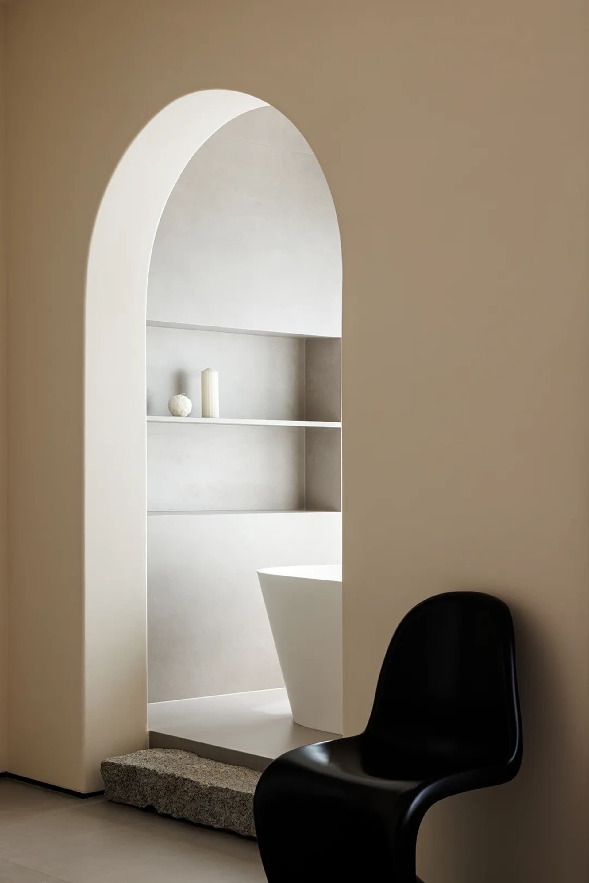A minimalist bathroom with a white bathtub and a black chair against a beige wall, framed by an arched doorway and a stone st