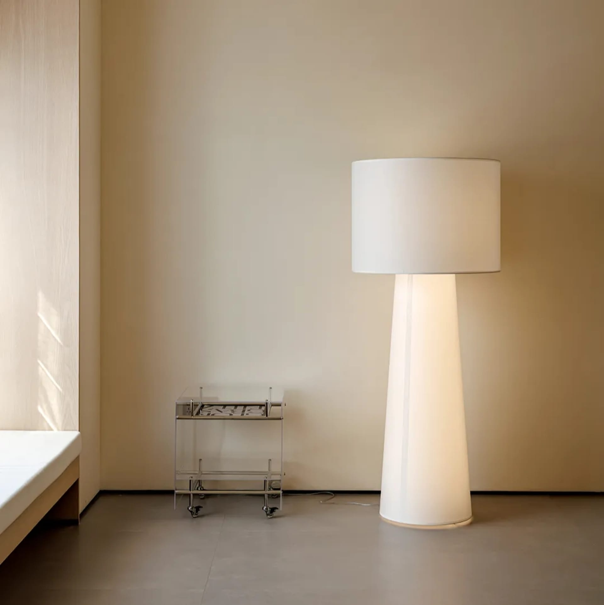 A minimalist bedroom with a large white floor lamp, a metal side table, and a bed against a beige wall.
