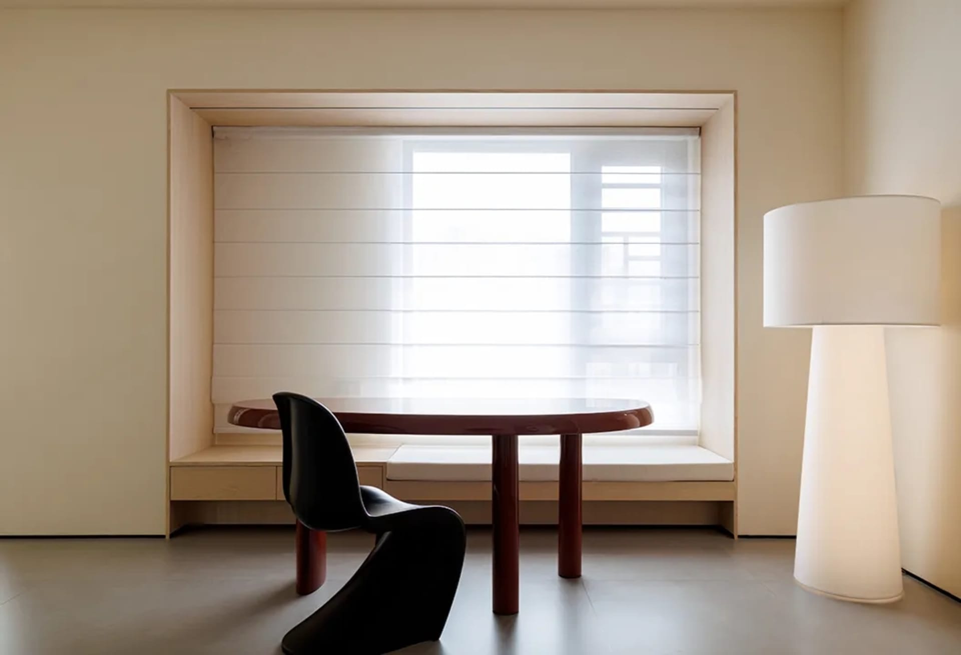 A modern home office featuring wood furniture, a round table, and a black chair, with a large window and a floor lamp.