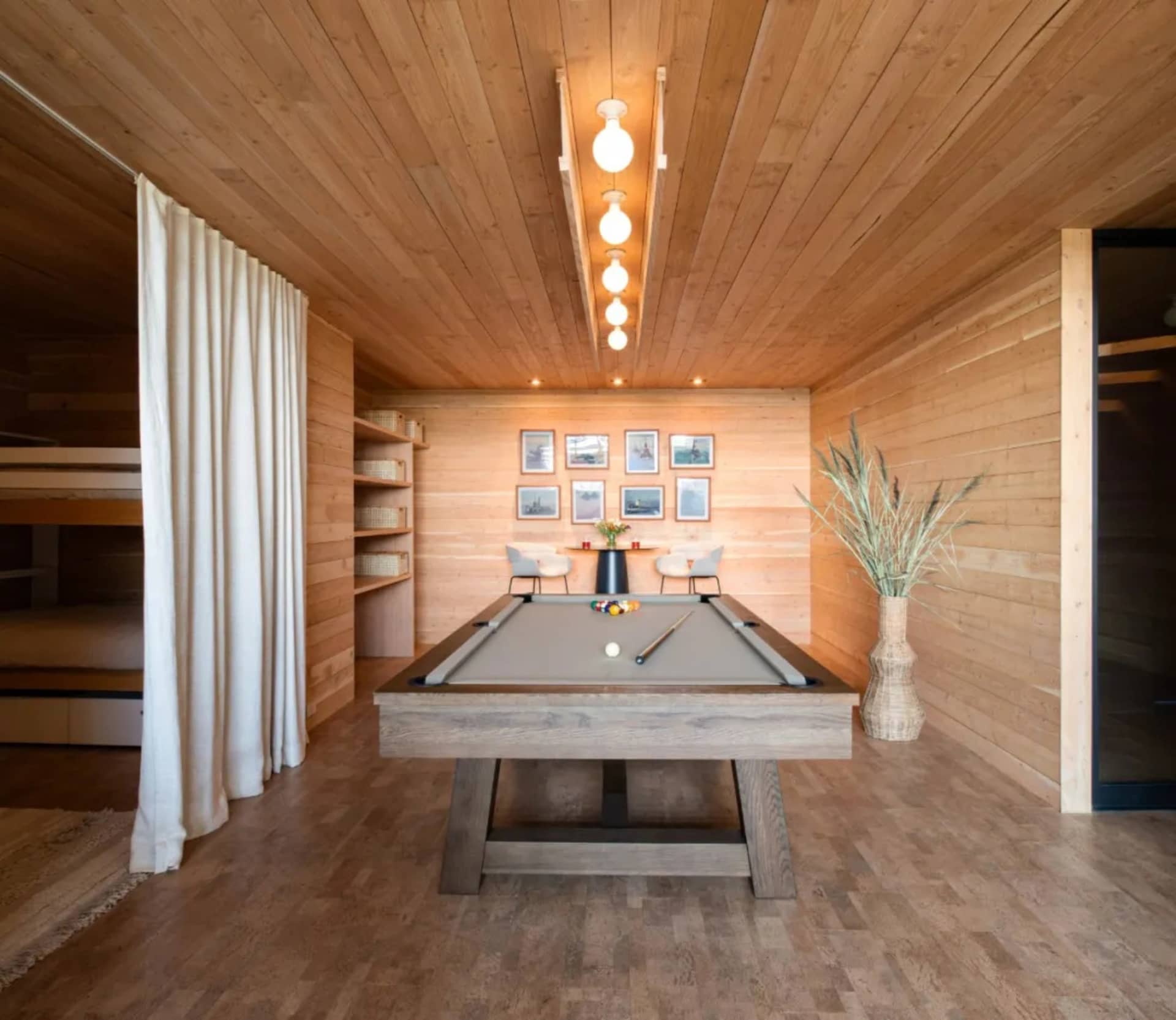 A modern minimalist dining room with a wooden wall, ceiling, and floor, featuring a gray pool table with balls and cues, whit