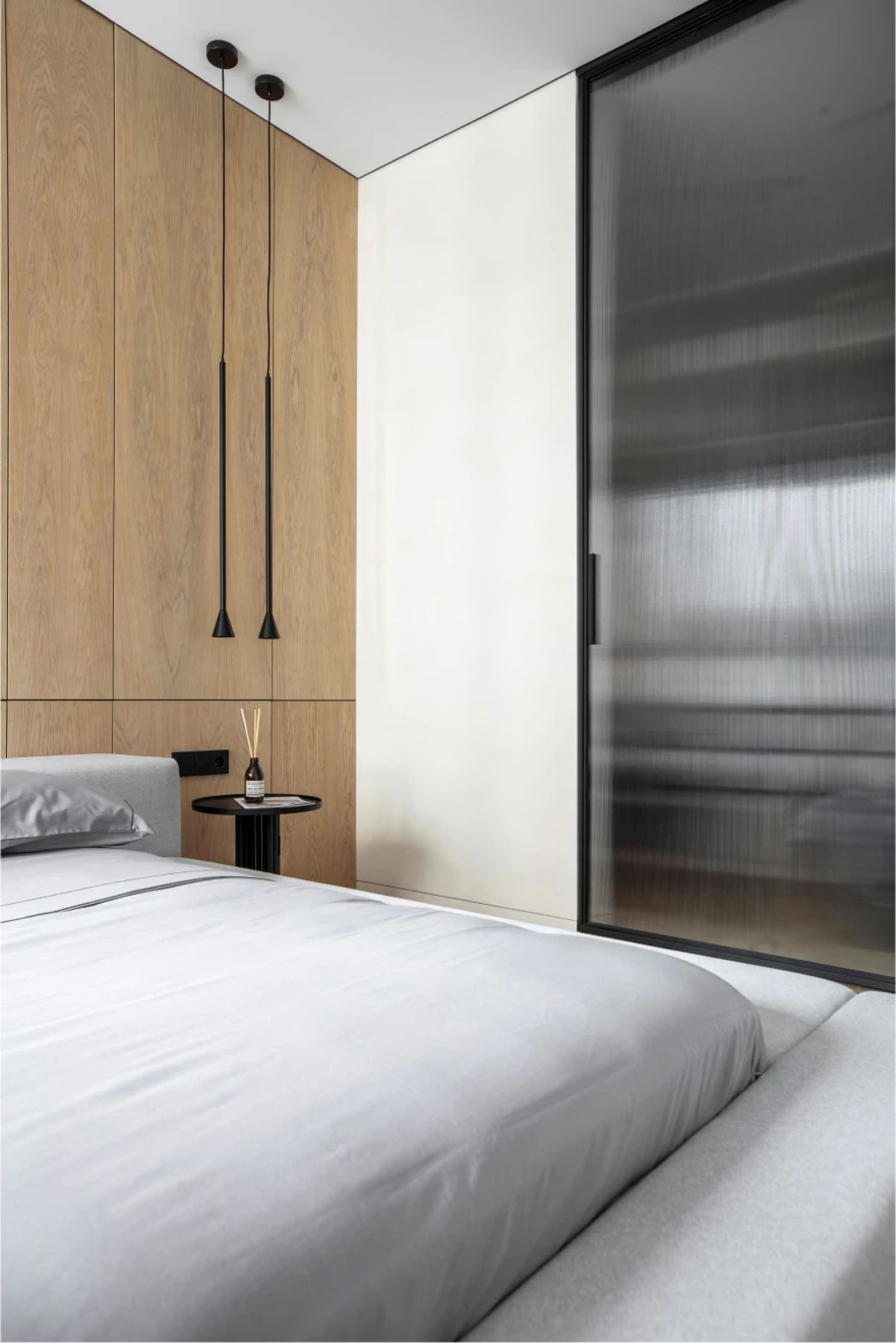 A sleek, modern bedroom featuring a bed with minimalist bedding, a small nightstand holding a decorative reed diffuser, and a