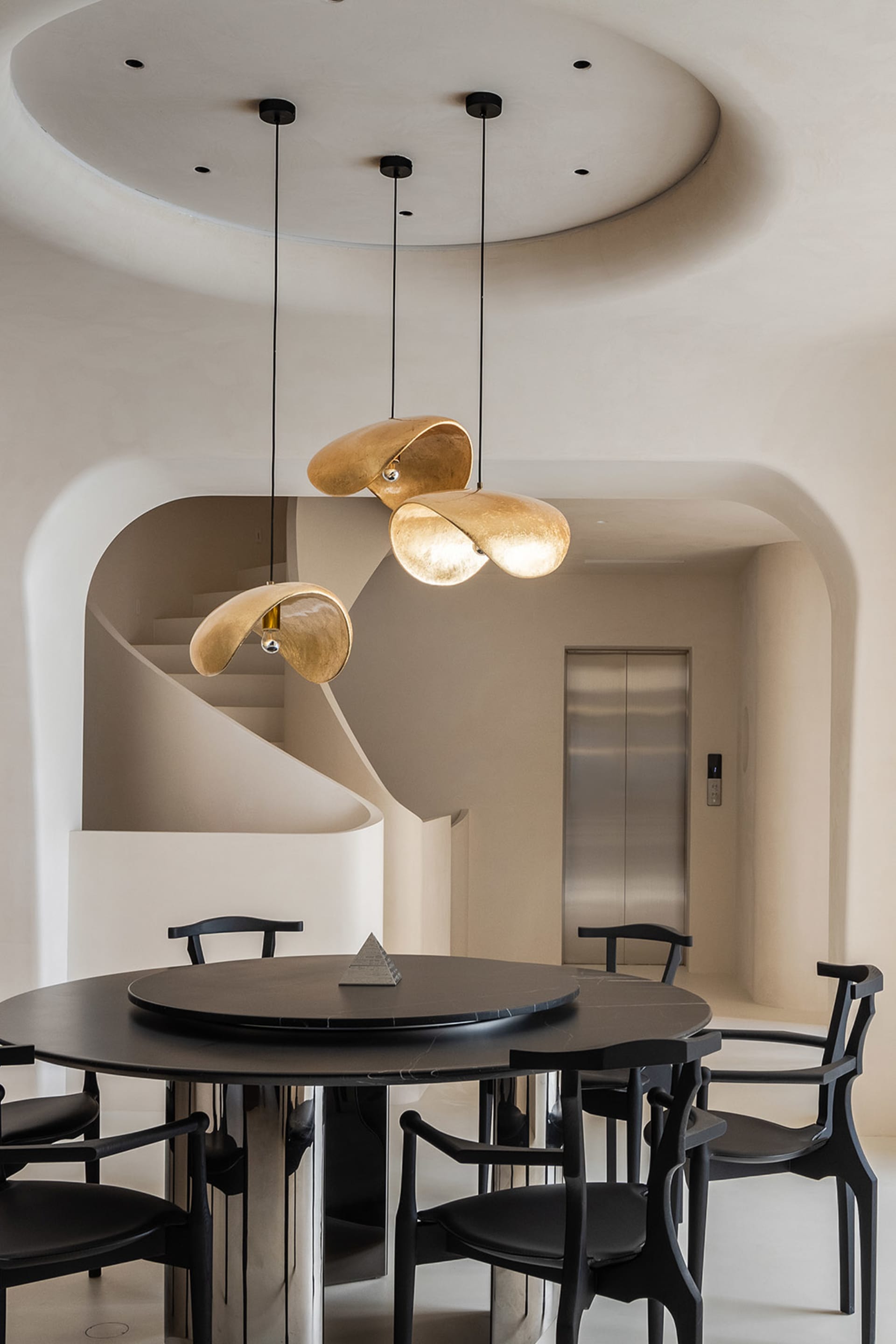 Modern dining room featuring a black, circular table with a central pyramid shape, black chairs, and three gold pendant light