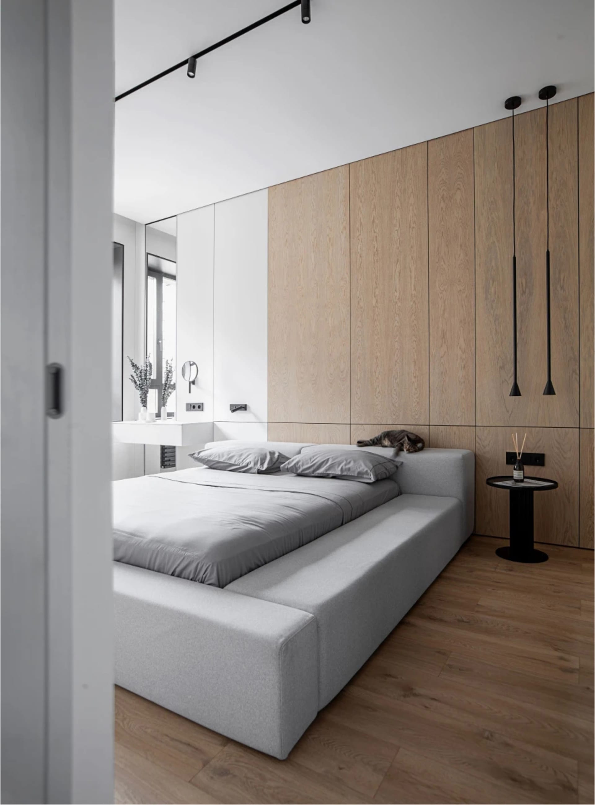 A minimalist bedroom with a gray bed on a platform, a wooden accent wall, and a white floating vanity with a black pendant li